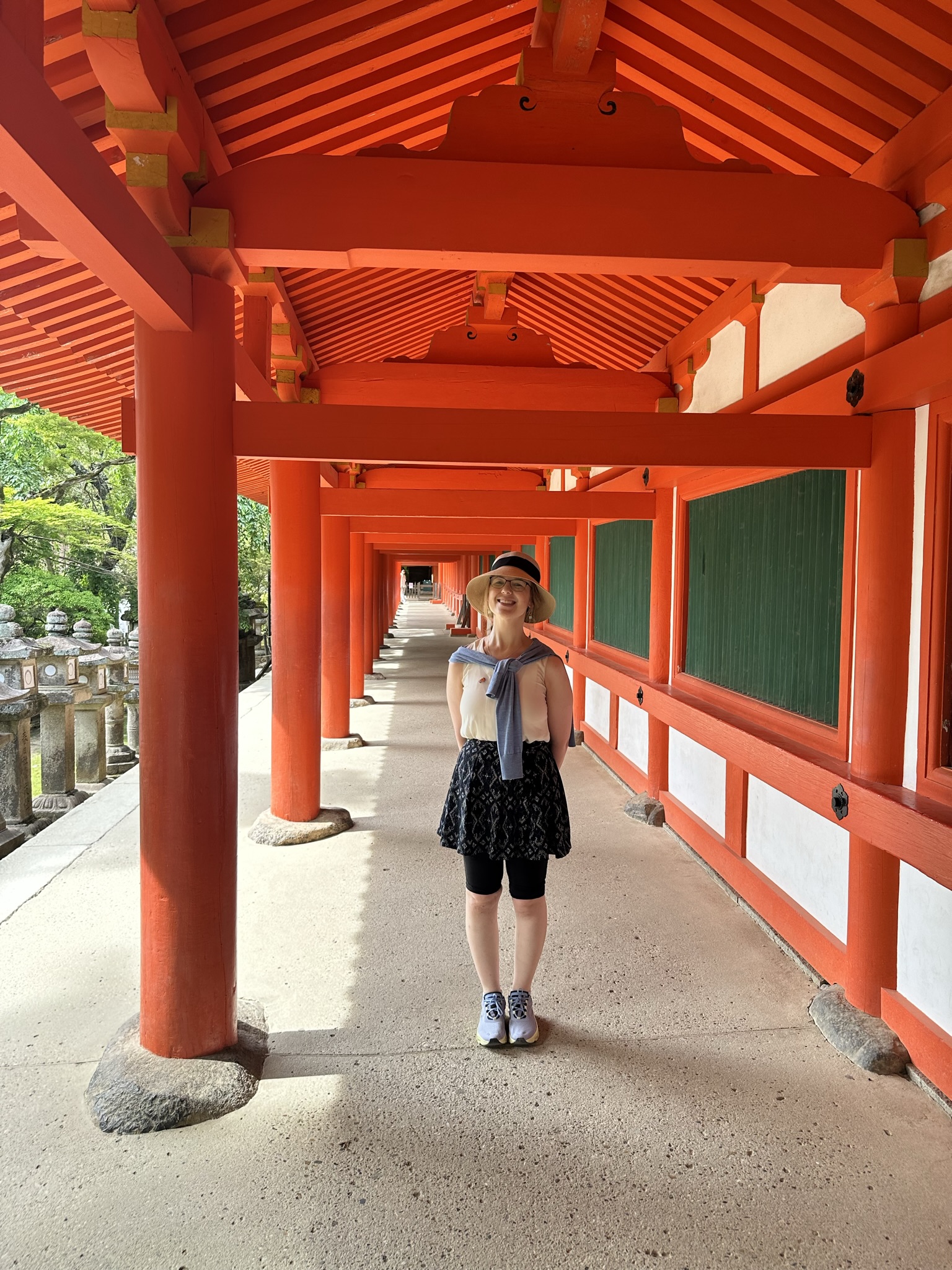 Mari-Alina Timoshchuk surrounded by red pillars at a Japanese temple in Nara Japan. She is wearing a large brimmed hat, blue skirt, and white blouse. She is smiling. There is a forest in the background. 
