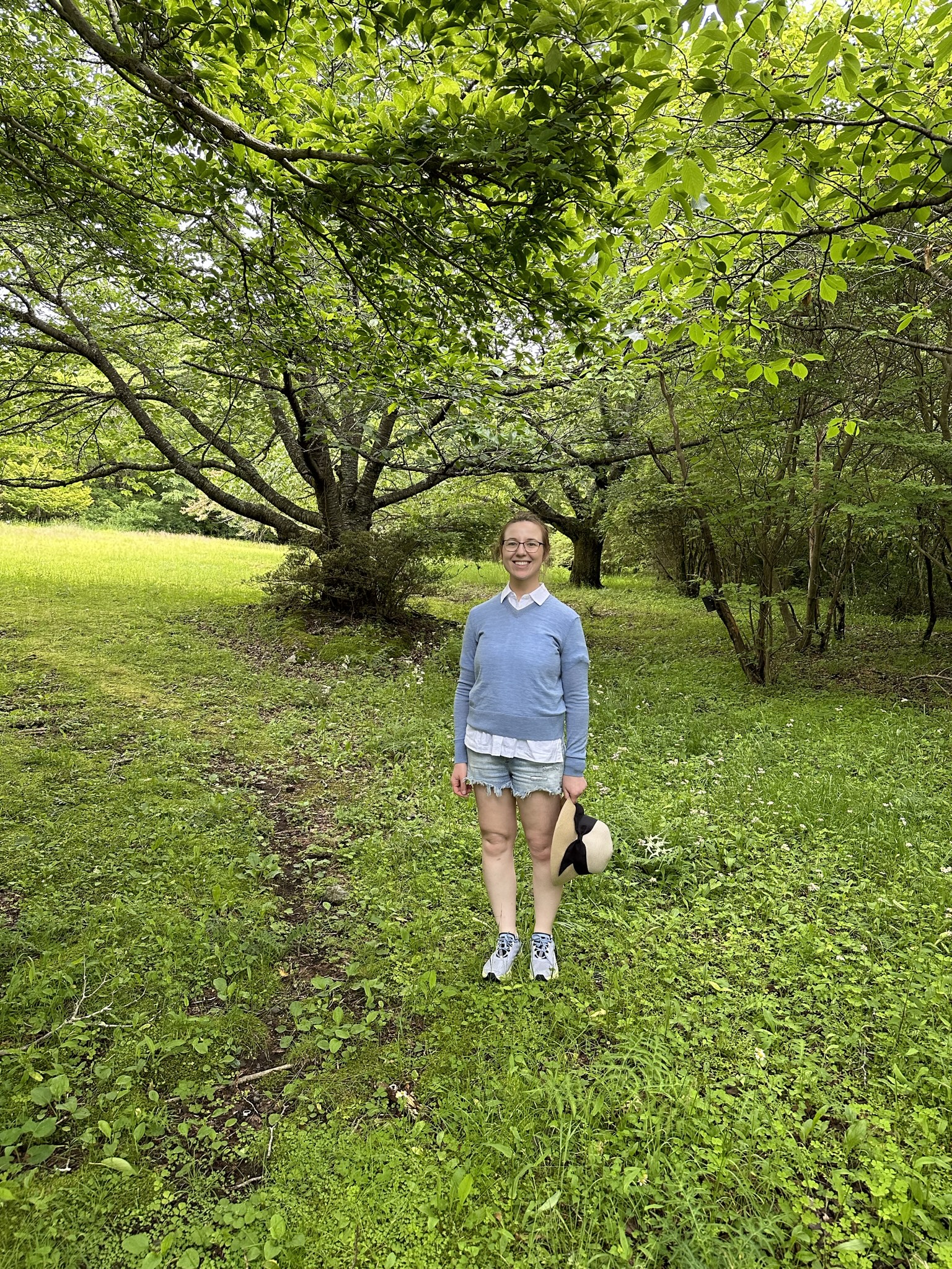 Mari-Alina Timoshchuk on a walk in Japan next to trees. She is wearing a blue sweater and shorts. She is holding a hat. She is smiling. 