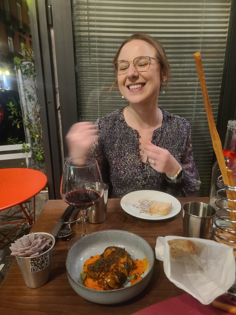 Mari-Alina Timoshchuk in Italy at a restaurant surrounded by food making gestures with her hand and smiling. She is in a colorful dress.