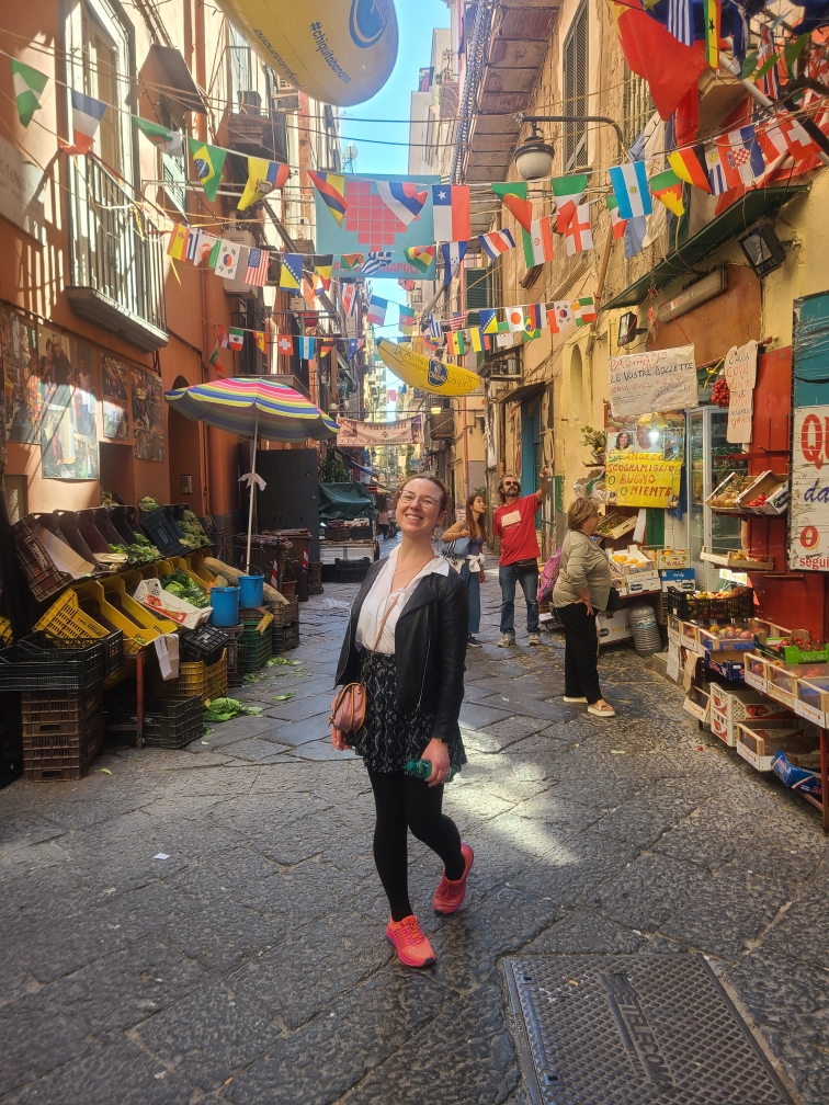 Mari-Alina Timoshchuk in front of a street market in Naples Italy. She is smiling and wearing pink shoes with a leather jacket. 