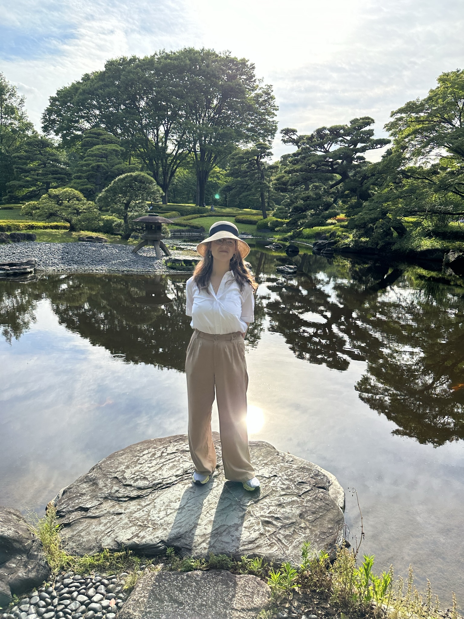 Mari-Alina Timoshchuk next to a lake in Tokyo standing on a rock with sunshine. She is wearing a wide brimmed hat, long brown pants, and a white blouse. She is smiling. 