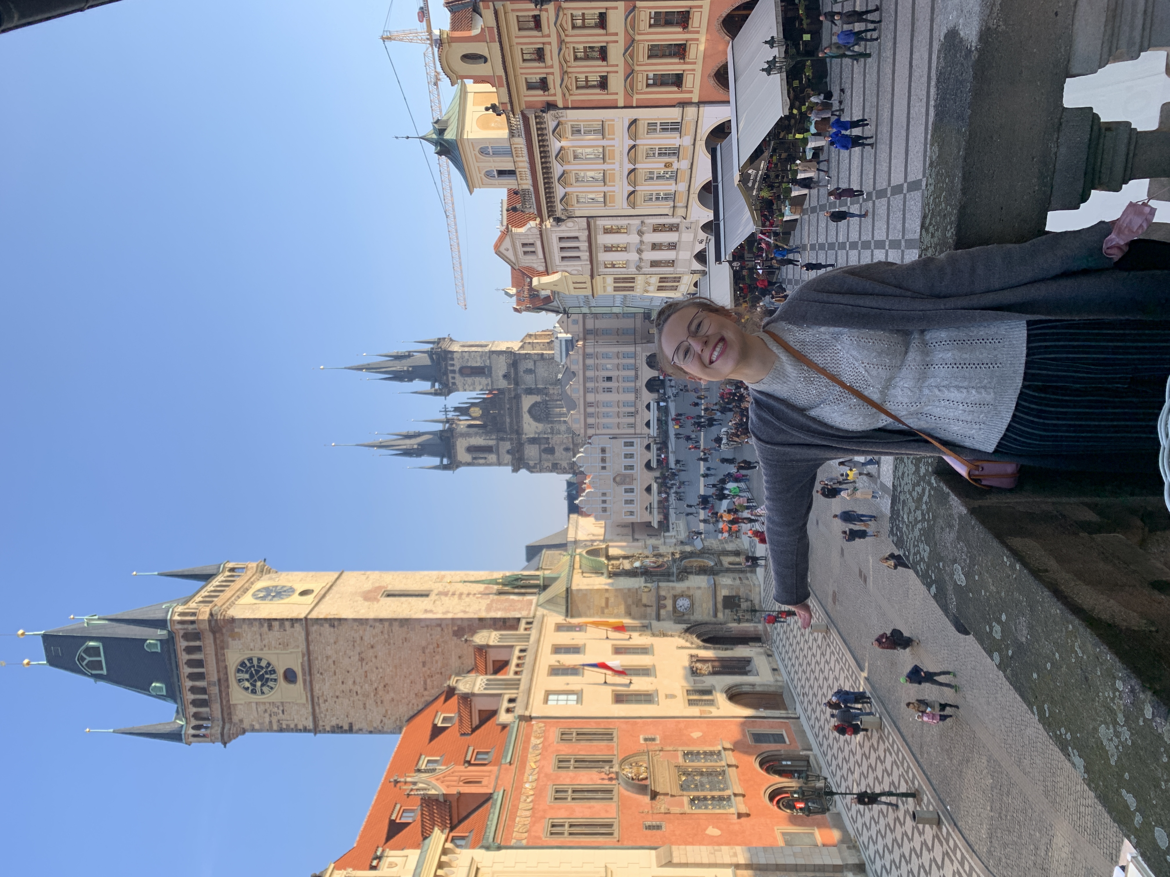 Mari-Alina Timoshchuk on a balcony in Prague overlooking Old Time Square. She has her arms spread and is smiling. 