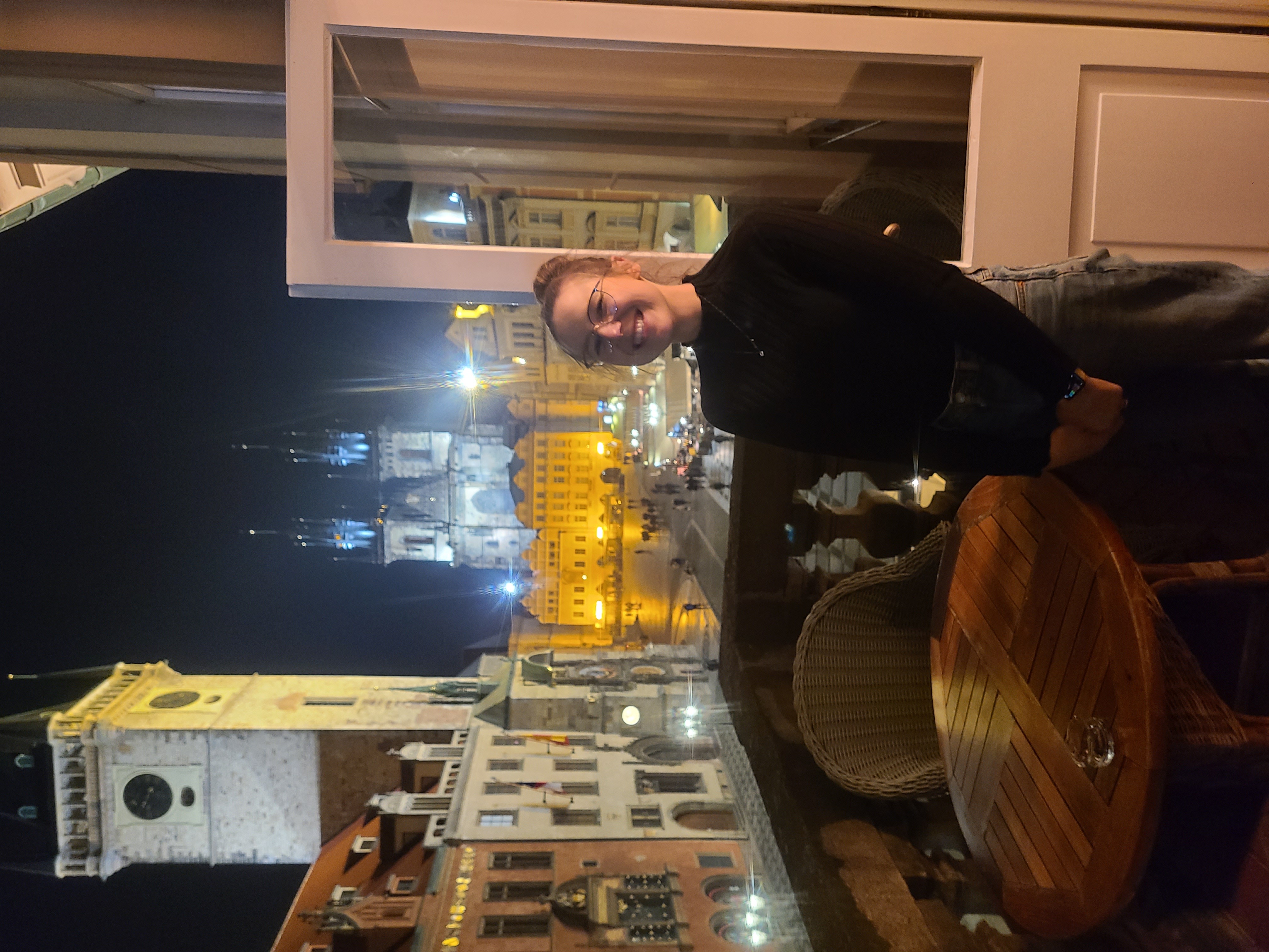 Mari-Alina Timoshchuk on a balcony in Prague at night with Old Time Square behind her. She is wearing a black turtle neck 