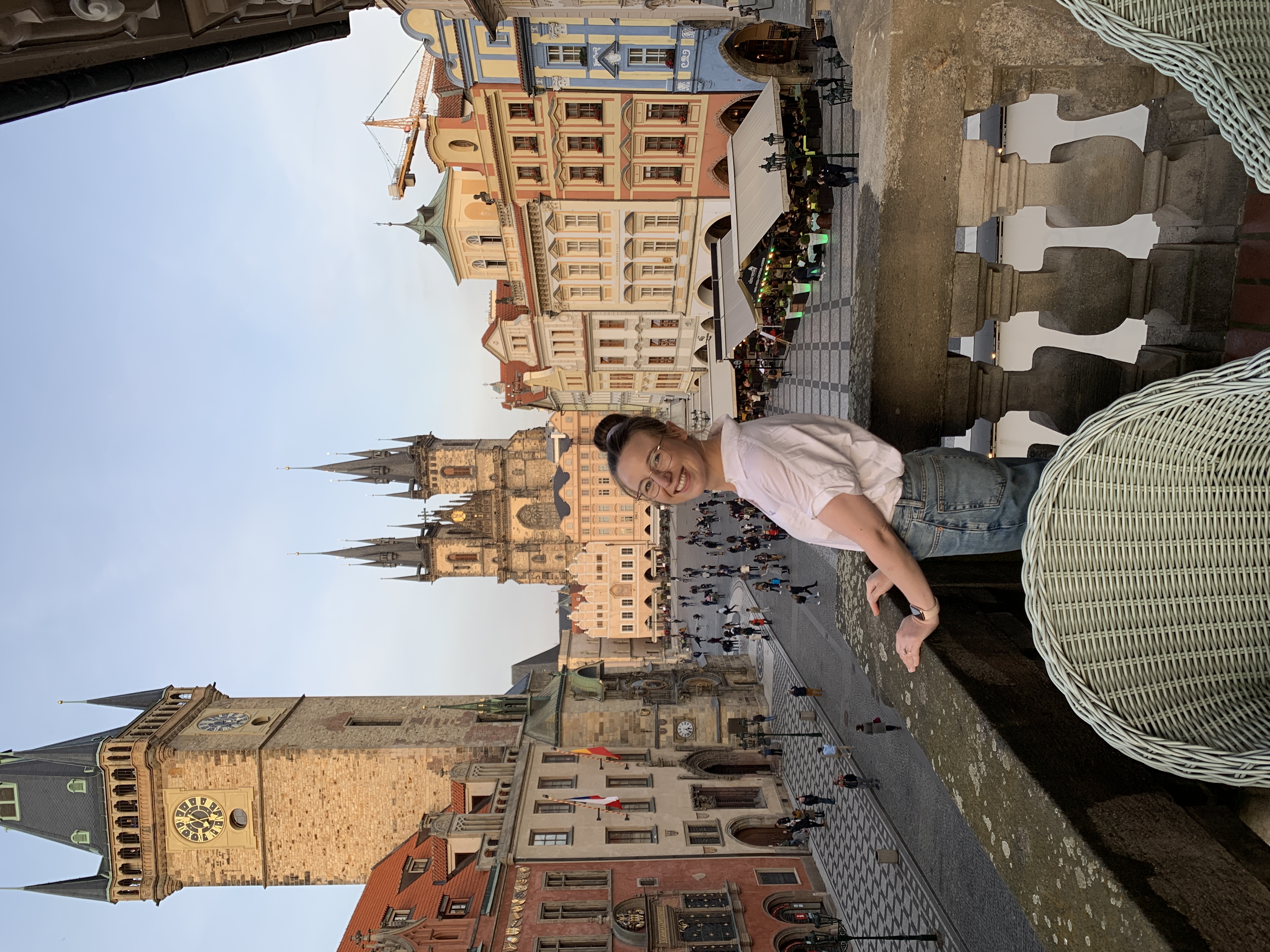 Mari-Alina Timoshchuk on a Prague balcony facing old town square. She is smiling and wearing jeans and a blouse. 