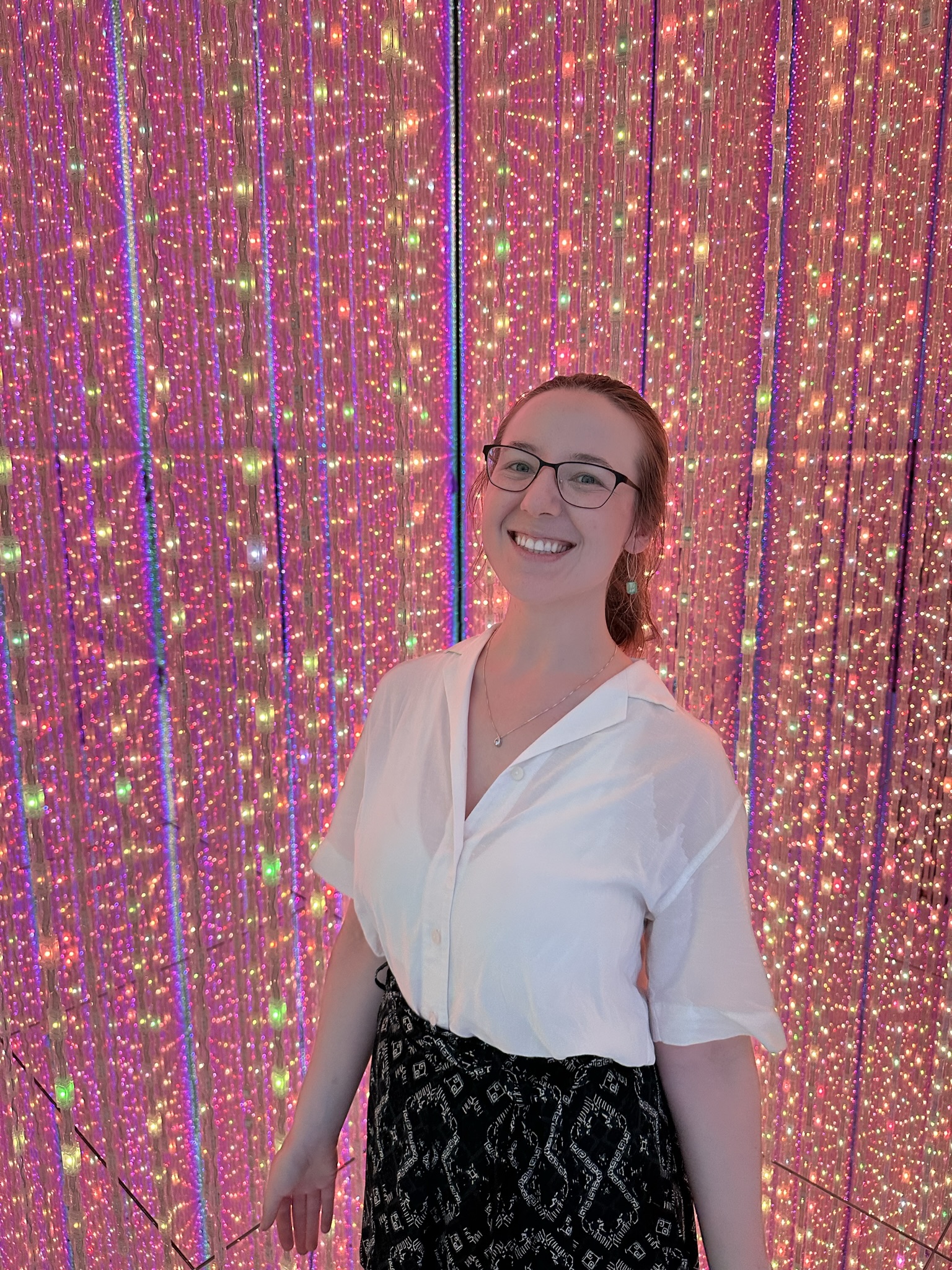 Mari-Alina Timoshchuk at teamLabs Planets in Tokyo next to a lot of pink lights at their infinite light exhibit. She is wearing a white blouse and blue skirt. She is smiling. 