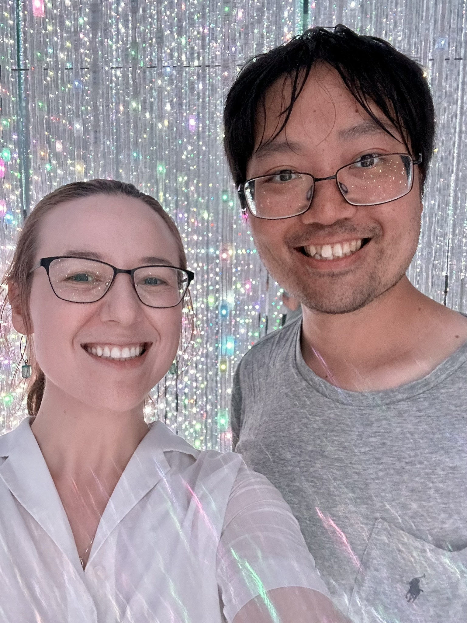 Mari-Alina Timoshchuk and Cassidy sung in teamLabs planets thing a selfie at the infinite light exhibit with silver lights in the background. They are both smiling. 