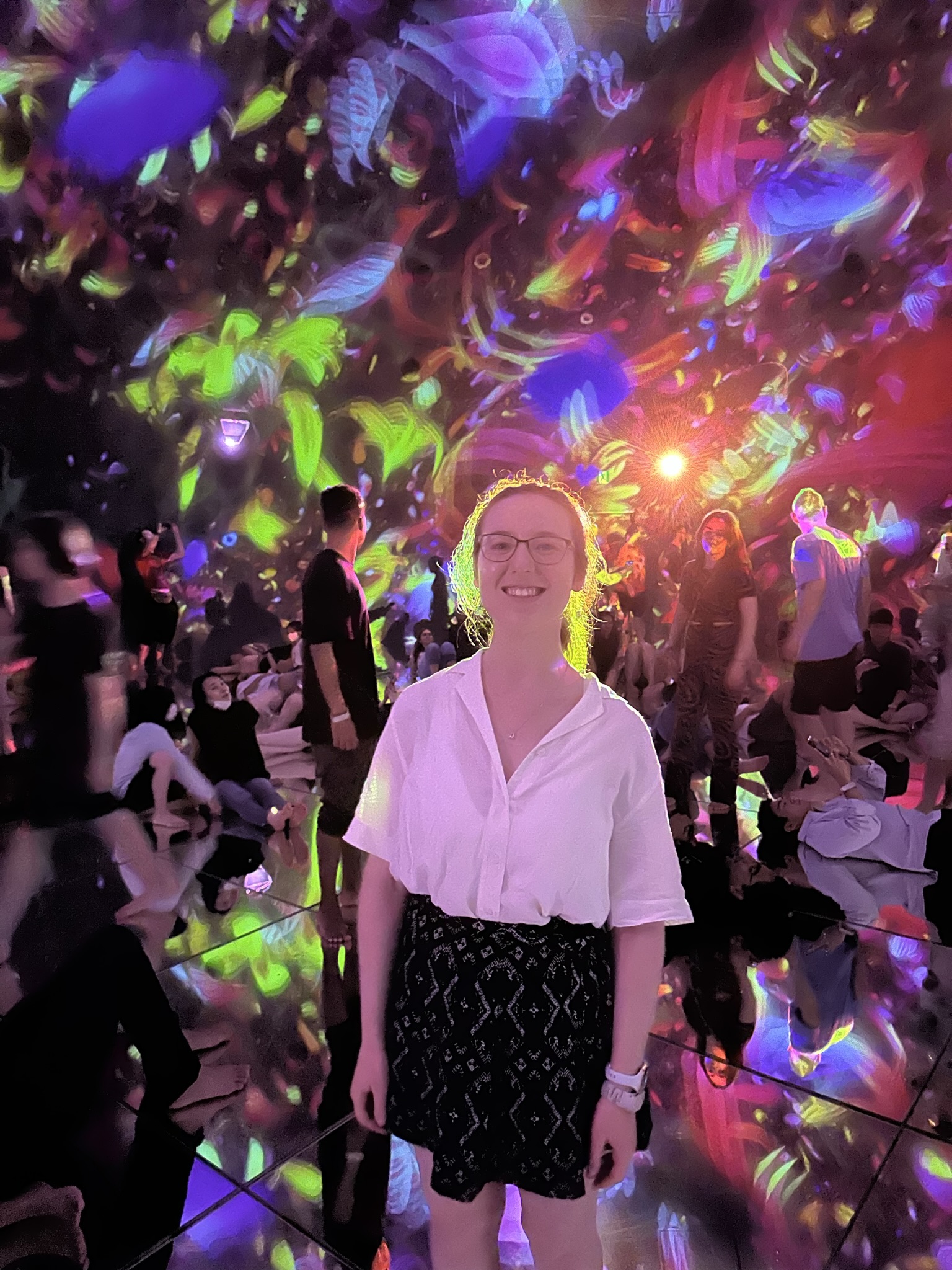 Mari-Alina Timoshchuk at the Floating Universe of flowers exhibit at teamLabs Planets in Tokyo. The background has many flowers in it. She is wearing a white blouse and blue skirt. She is smiling. 