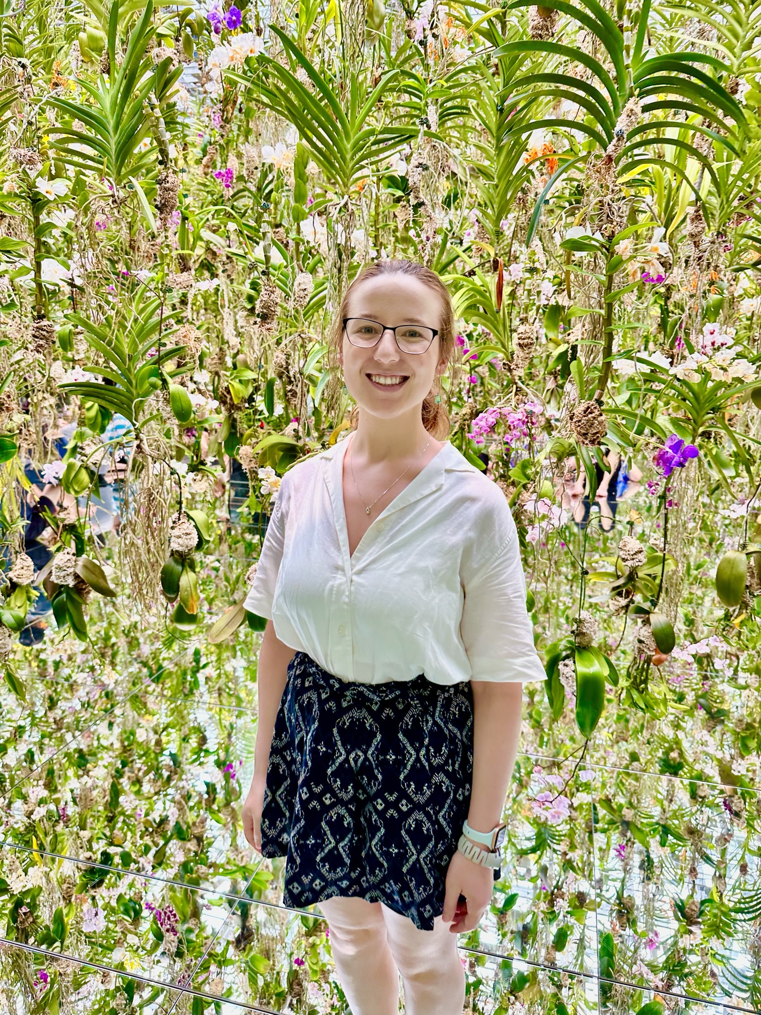 Mari-Alina Timoshchuk surrounded by Iris flowers in an art exhibit in Tokyo Japan. She is wearing a white blouse and a blue skirt. She is smiling. 