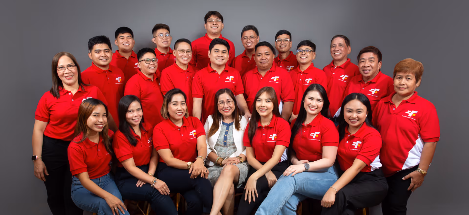 Group photo of 22 people, all wearing matching red collared shirts with a logo featuring stylized white and yellow letters. Most individuals are smiling, posed in three rows against a plain gray backdrop, suggesting a corporate or team setting. One woman in the front center stands out in a patterned dress and white blazer, likely a leader or executive.