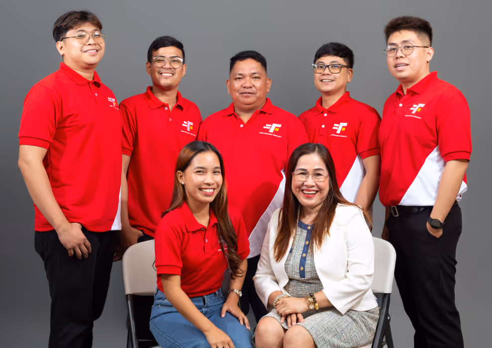 Group photo of seven people in red collared shirts with a company logo, posed in front of a gray backdrop. Two women are seated at the front, smiling—one in casual jeans and the other in a white blazer and tweed dress—while five men stand behind them in matching uniforms, suggesting a collaborative and approachable team.