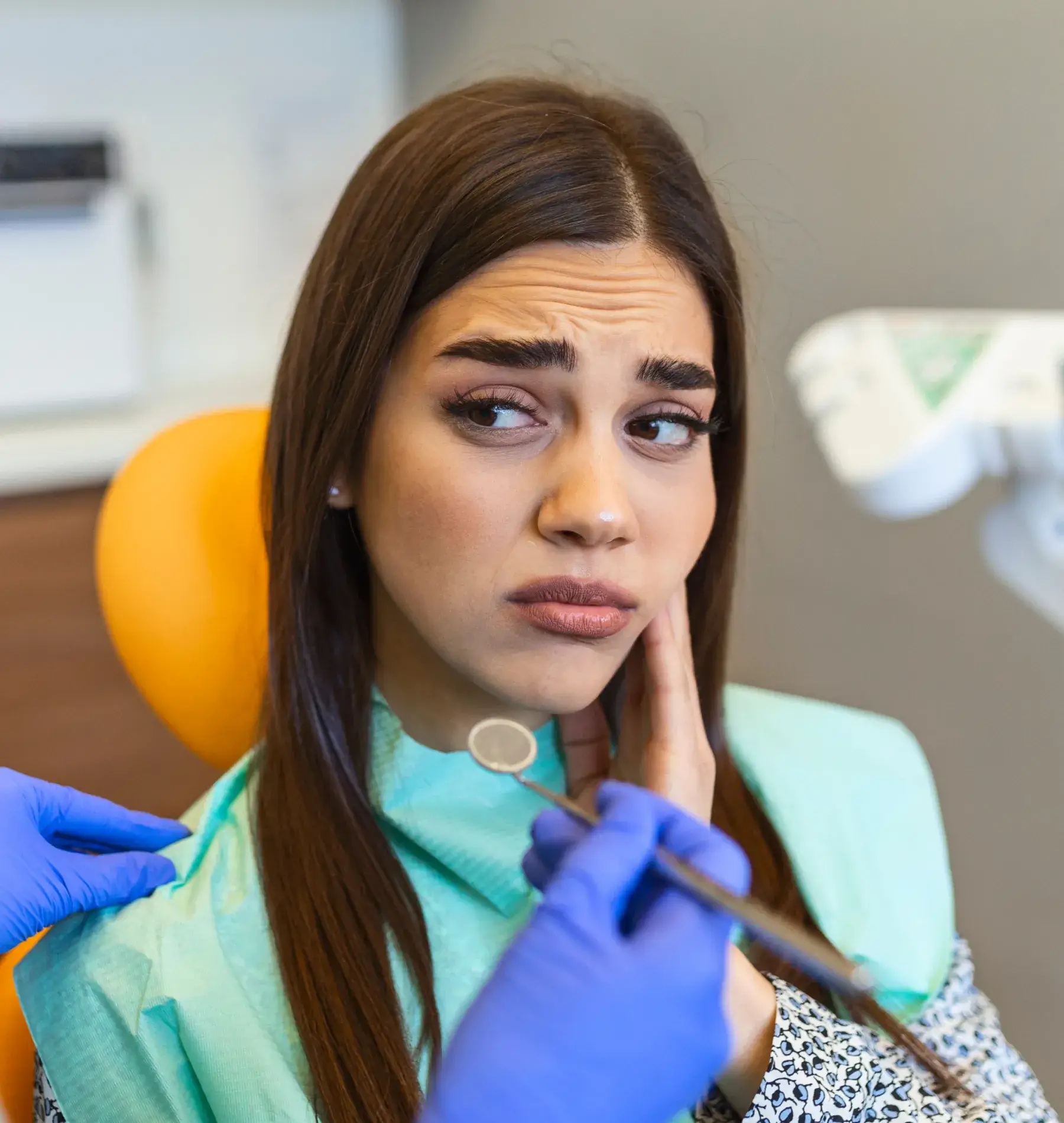 A woman looks worried at the dentist while a dental professional holds tools near her mouth.