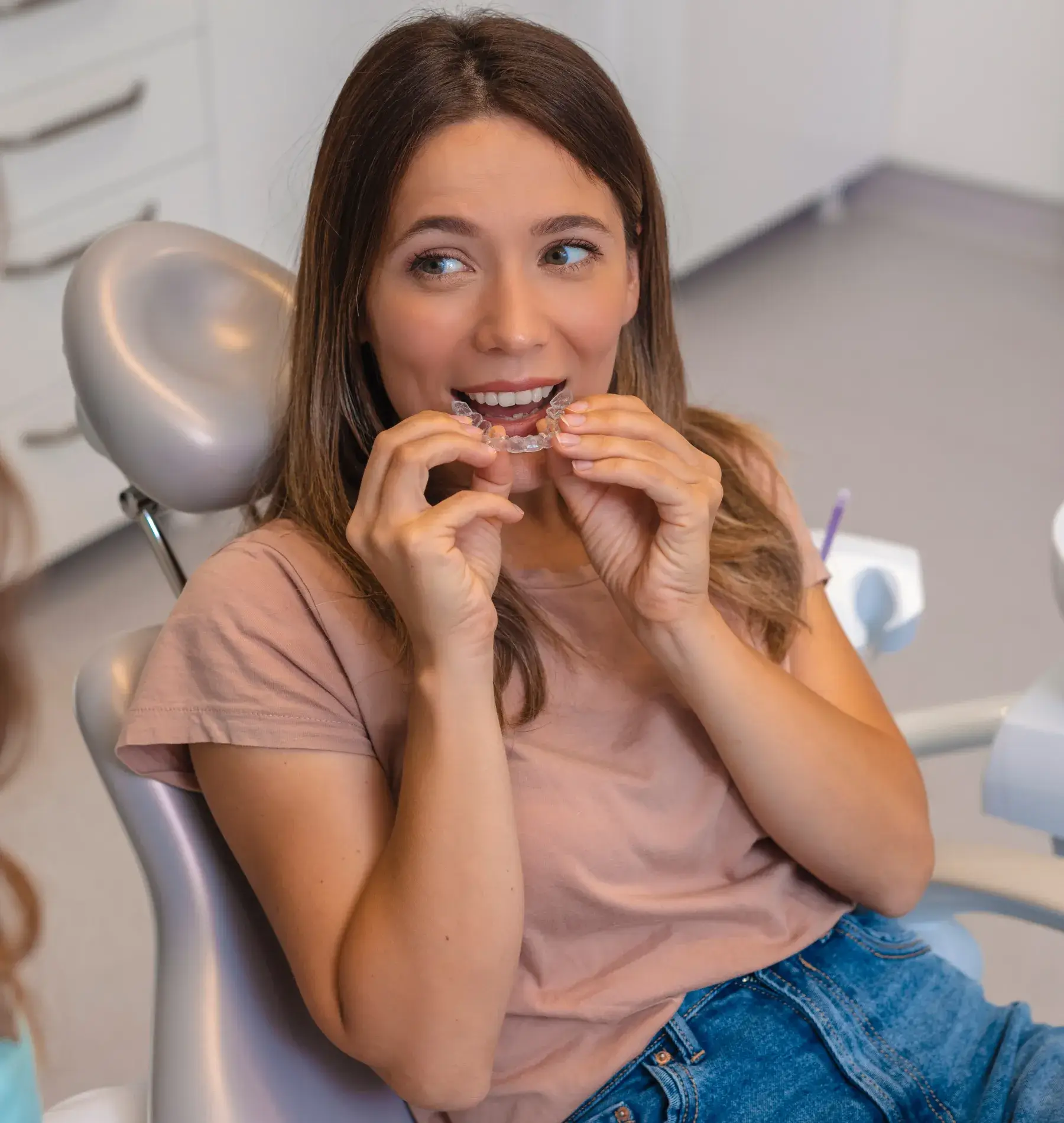 A woman sitting in a dental chair holds an orthodontic retainer in her hands, preparing to insert it.