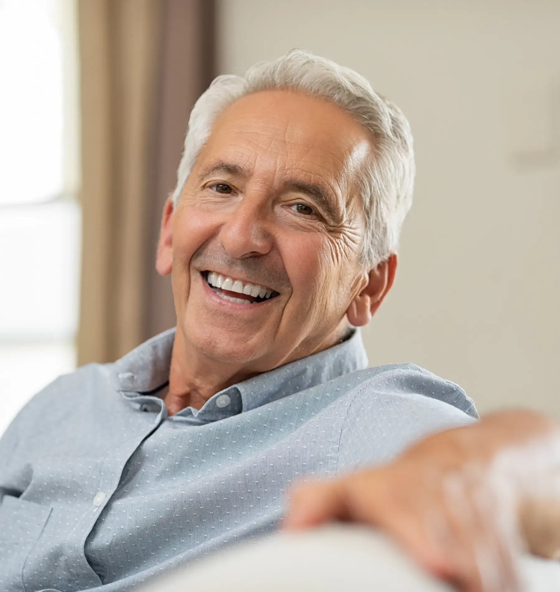 Older man smiling while sitting on a couch in a softly lit room.