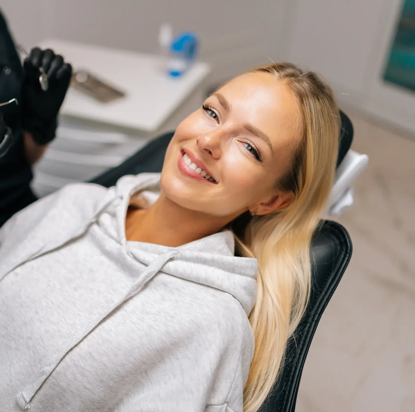 Smiling blonde woman in a gray hoodie reclining in a dental chair during a dental visit.