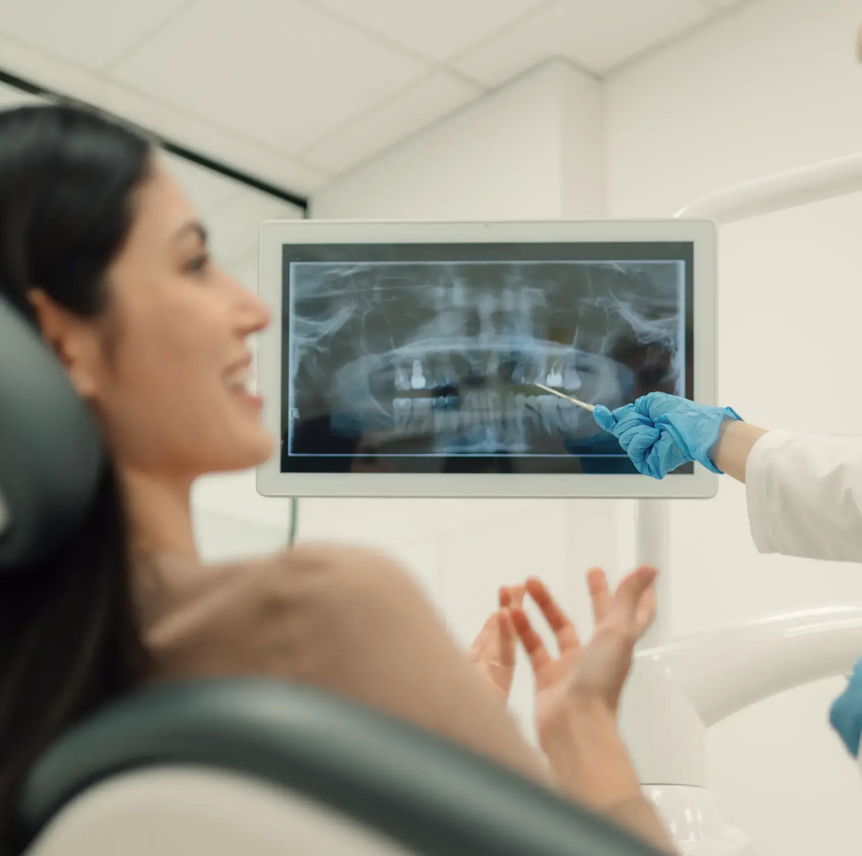 Dentist wearing blue gloves pointing at a dental X-ray on a screen while a female patient smiles in a dental chair.