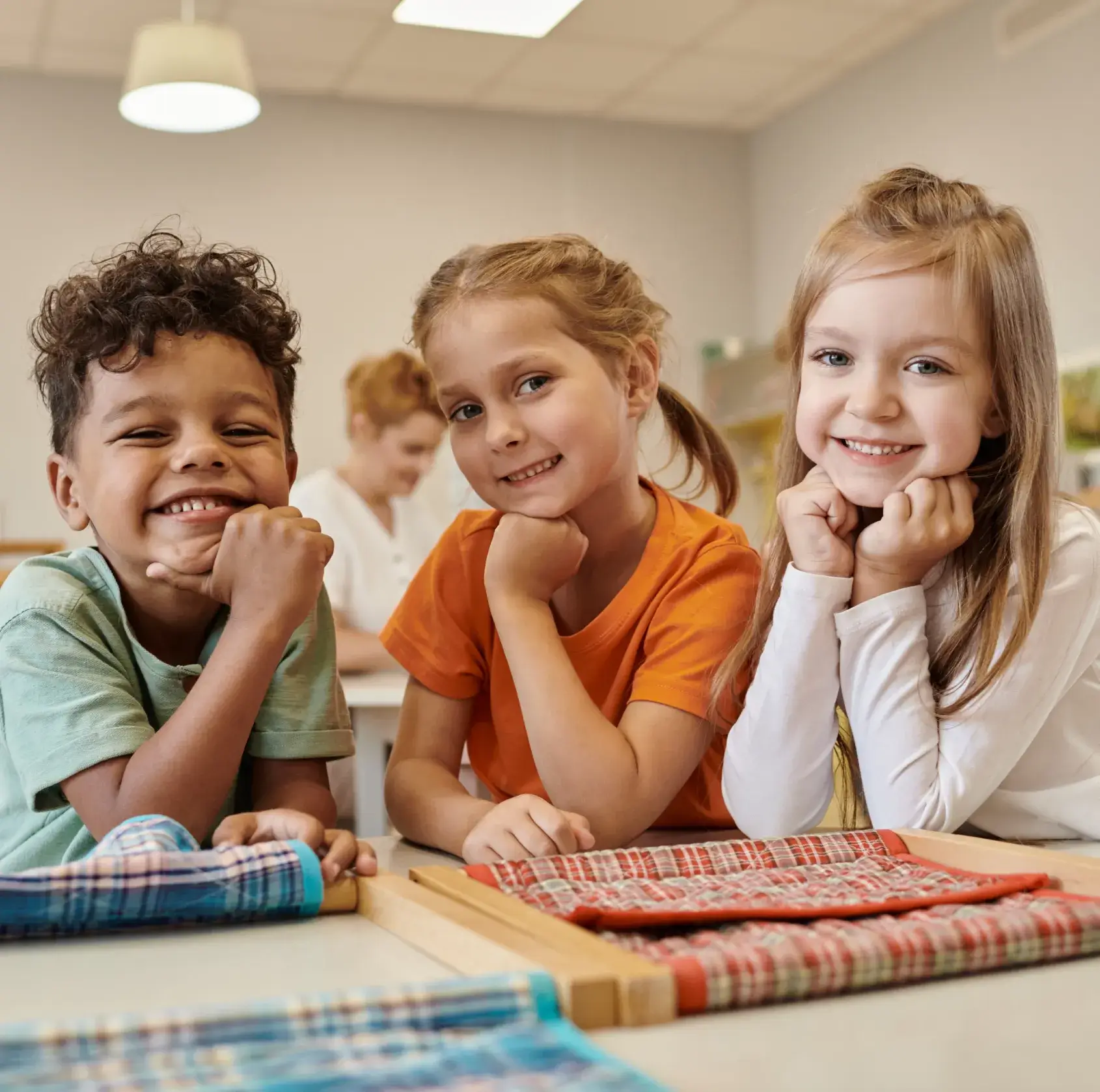 Three smiling children sitting at a table with checkered fabric activity, resting their chins on their hands.