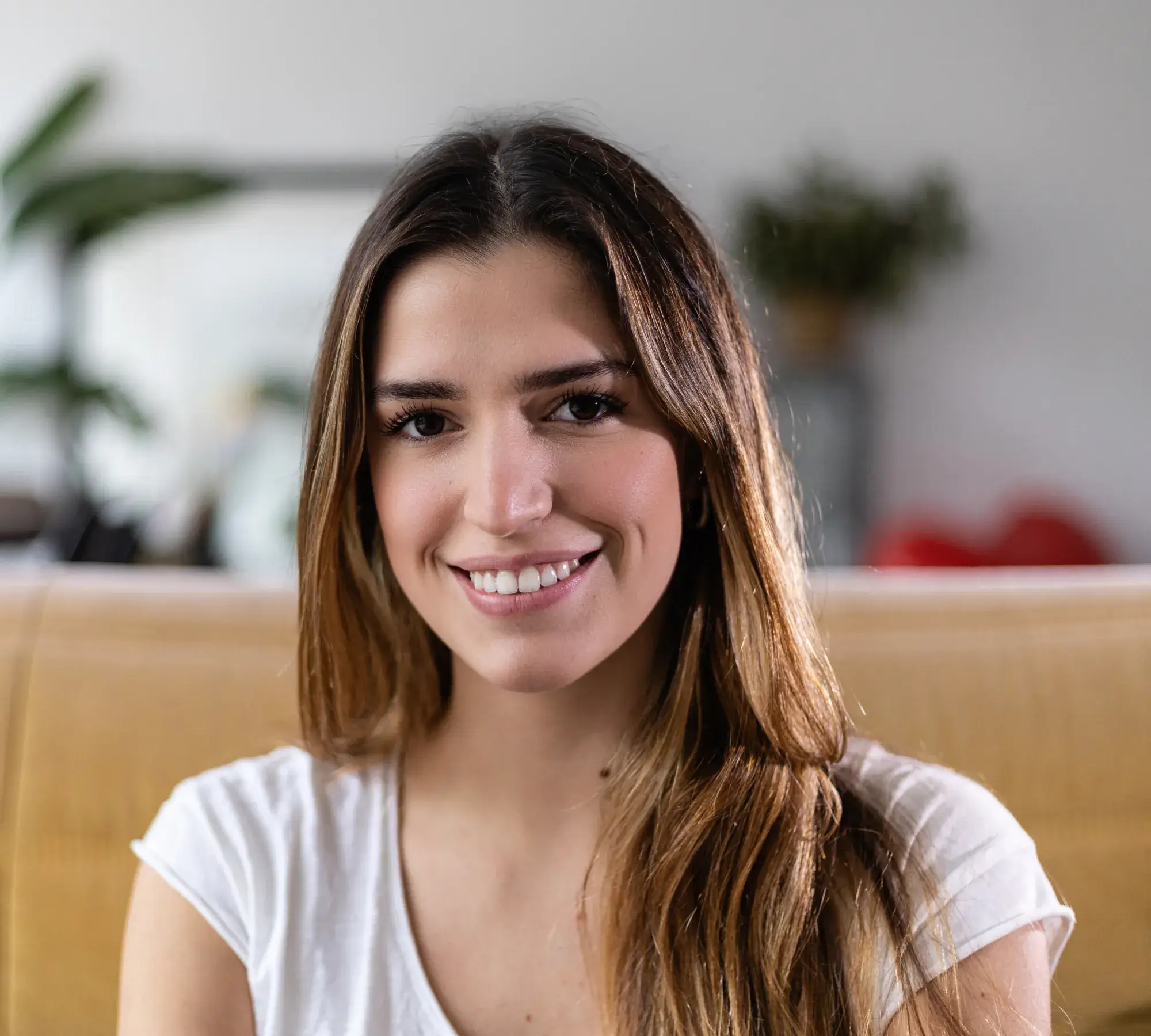 Smiling young woman with long brown hair wearing a white top sitting on a beige couch in a softly blurred room.