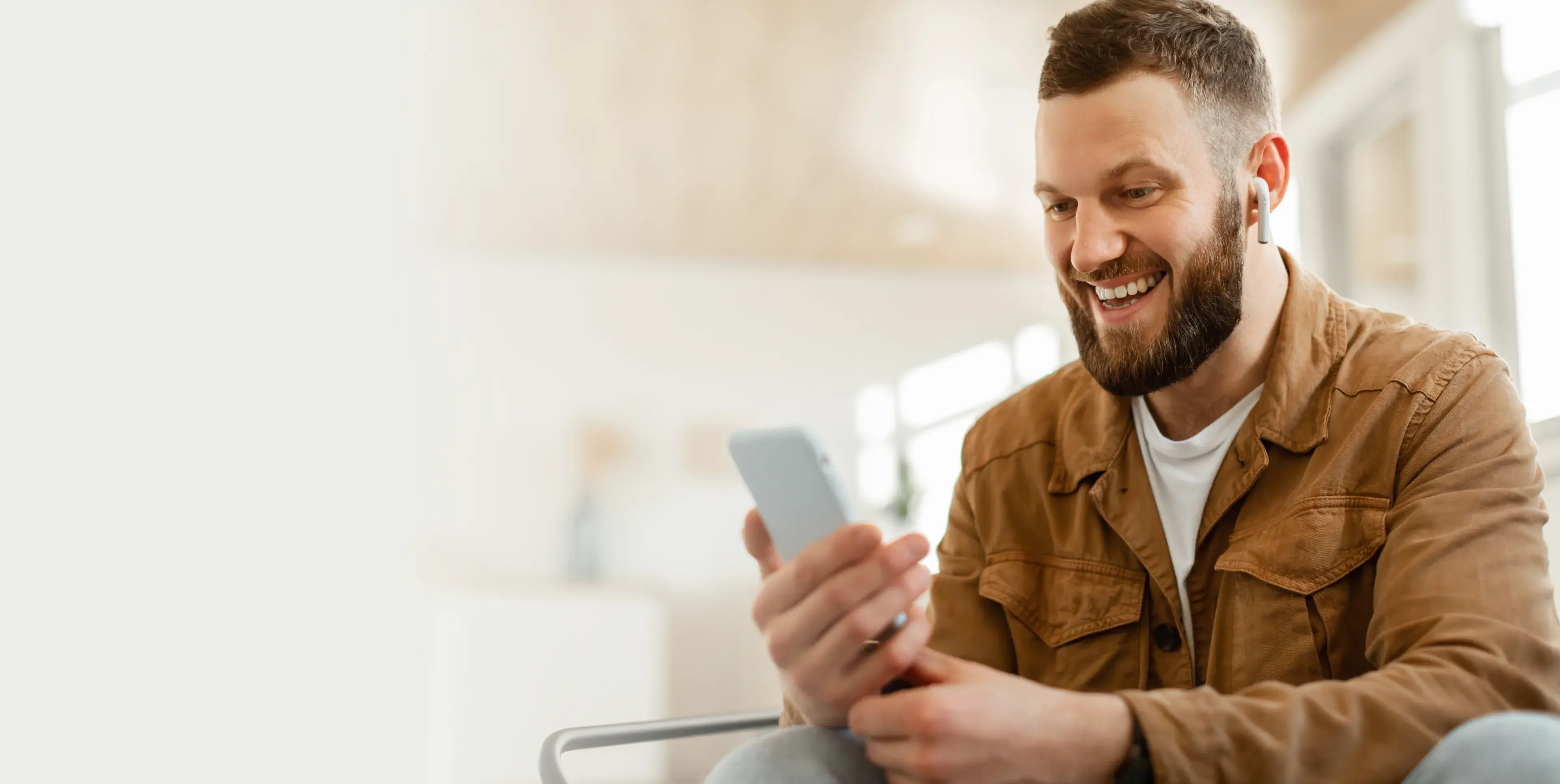 Smiling bearded man wearing earphones and a brown jacket looking at his smartphone indoors.