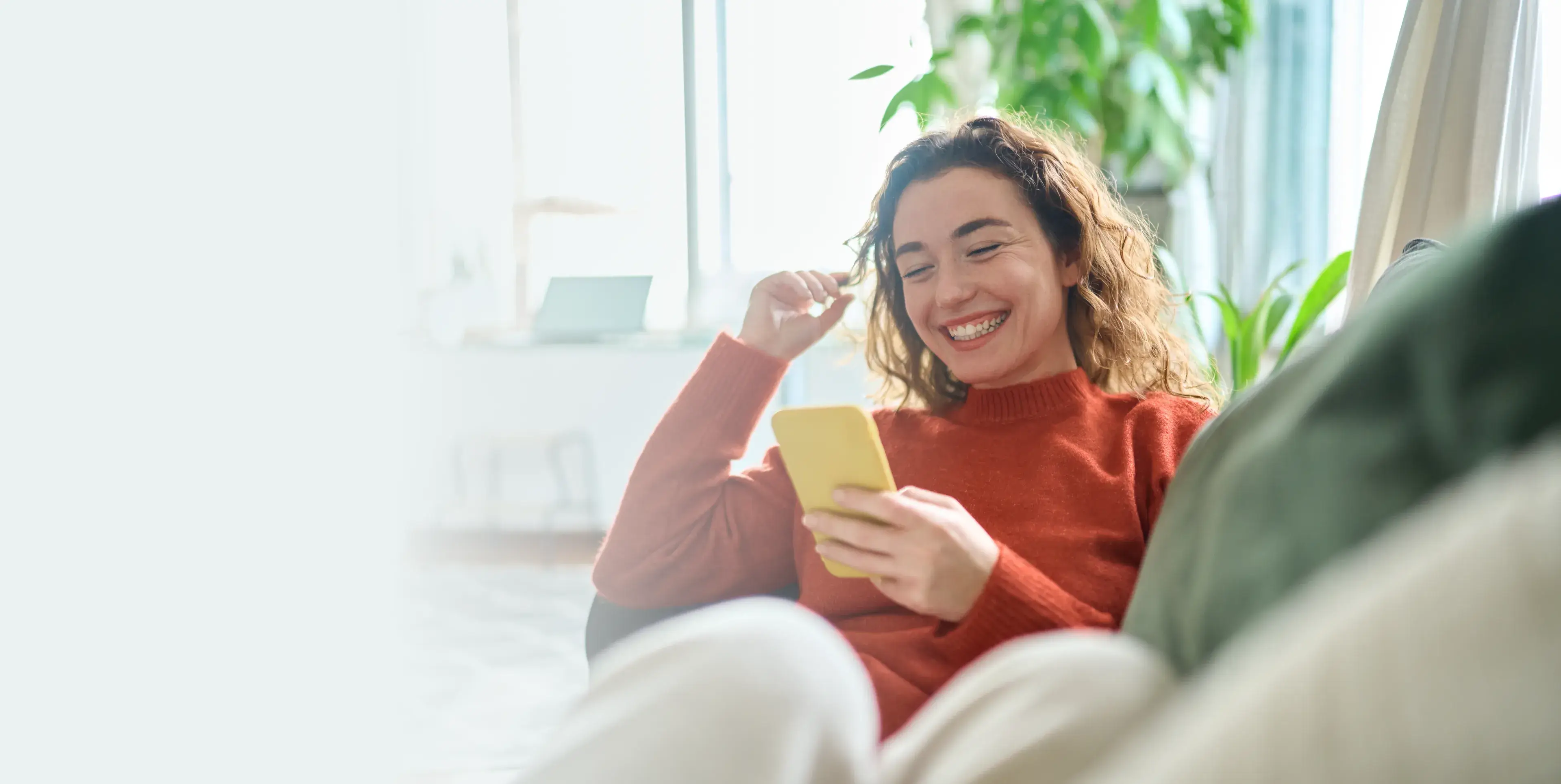 Smiling woman in an orange sweater sitting on a couch while looking at a yellow smartphone.