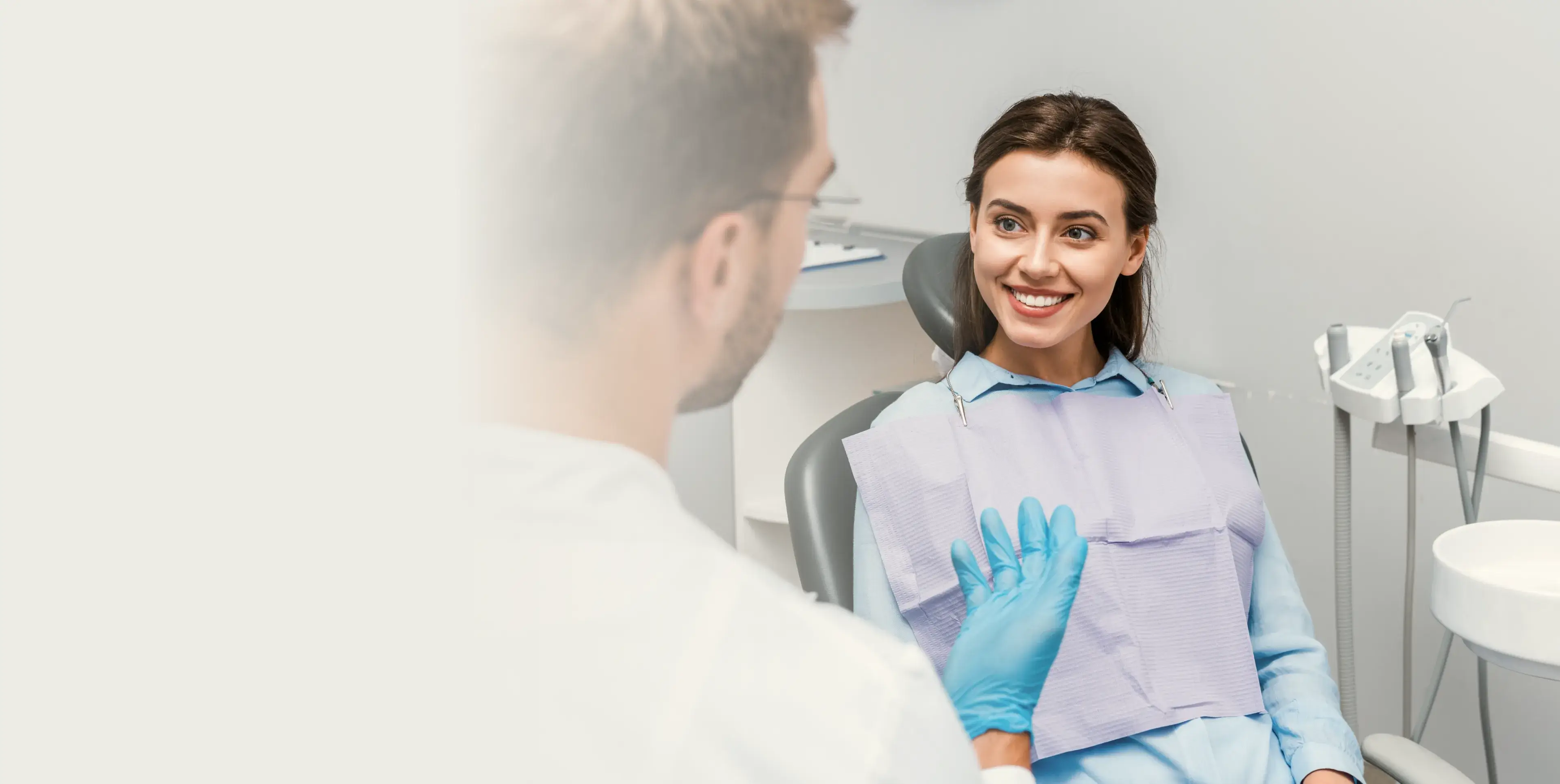 Smiling woman sitting in dental chair talking to a dentist wearing blue gloves in a dental office.