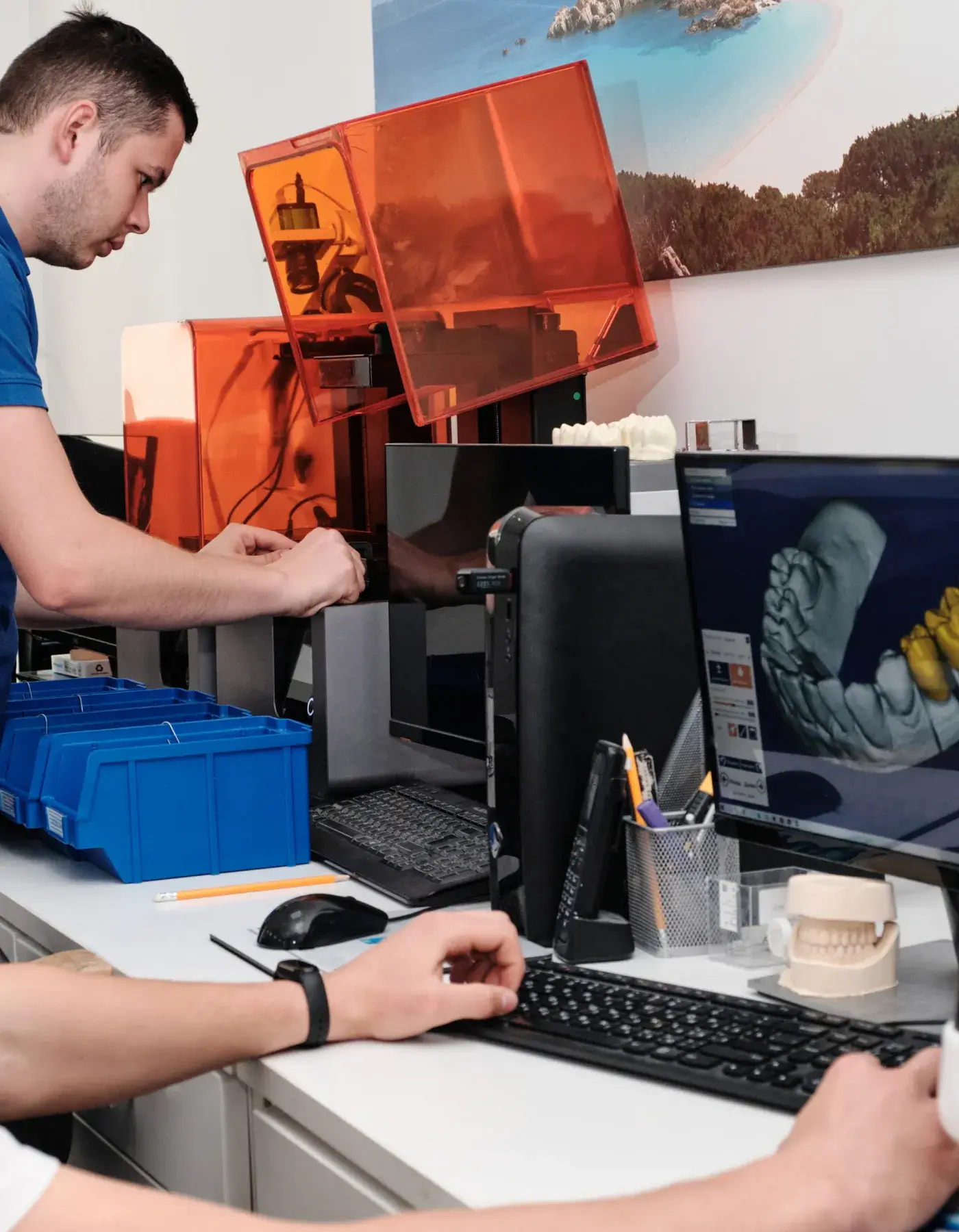 A man working with a 3D printer and computer showing a digital dental model on screen in a dental lab.