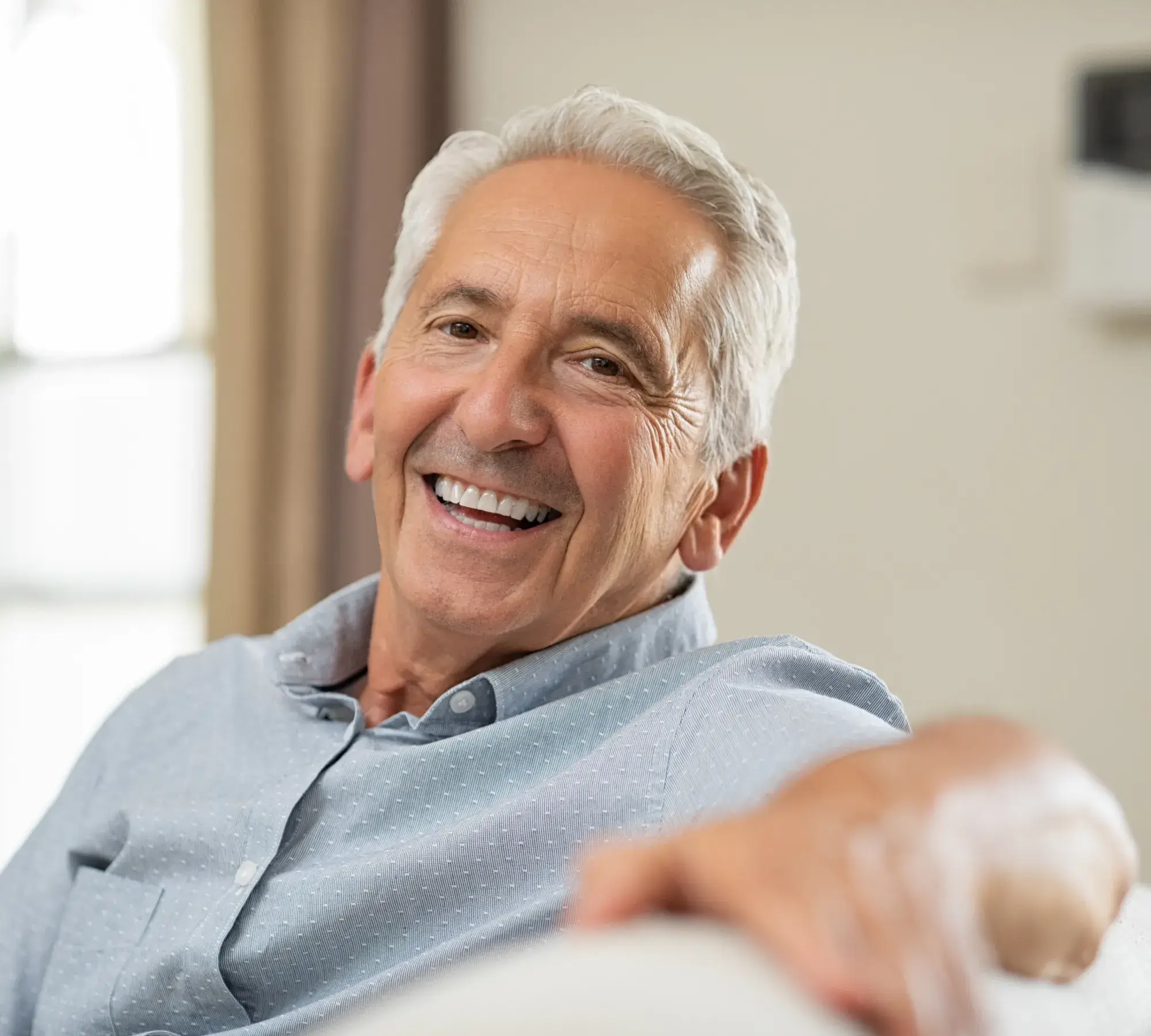 Smiling elderly man with gray hair sitting comfortably on a couch indoors.