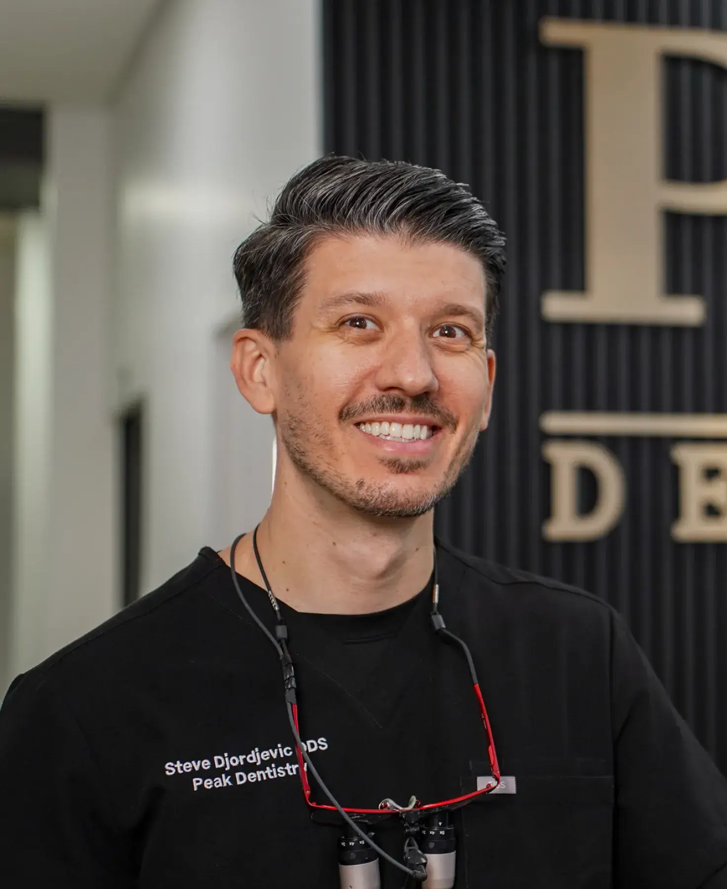 Smiling male dentist with short dark hair wearing black scrubs and dental loupes around his neck.