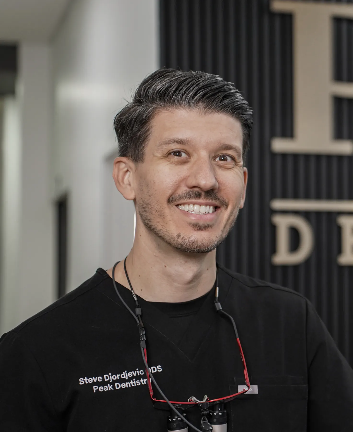 Smiling male dentist wearing black scrubs with dental magnifying glasses around his neck in a dental office.