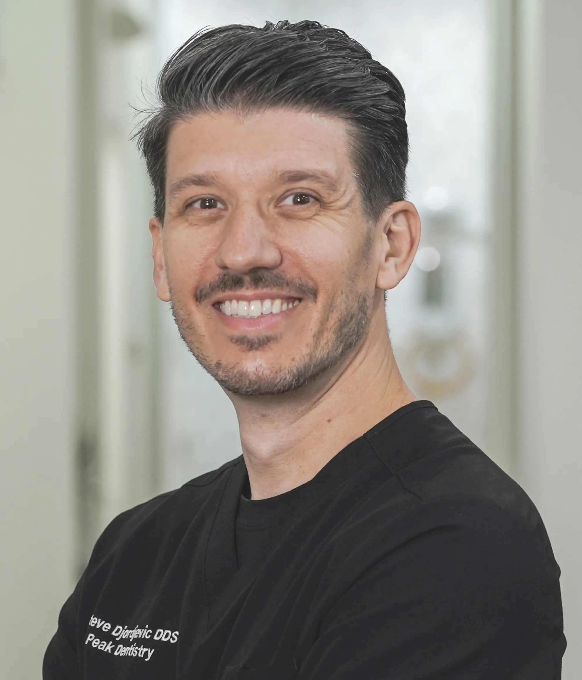 Smiling man with short dark hair and light facial stubble wearing a black shirt with dental practice name, standing indoors.