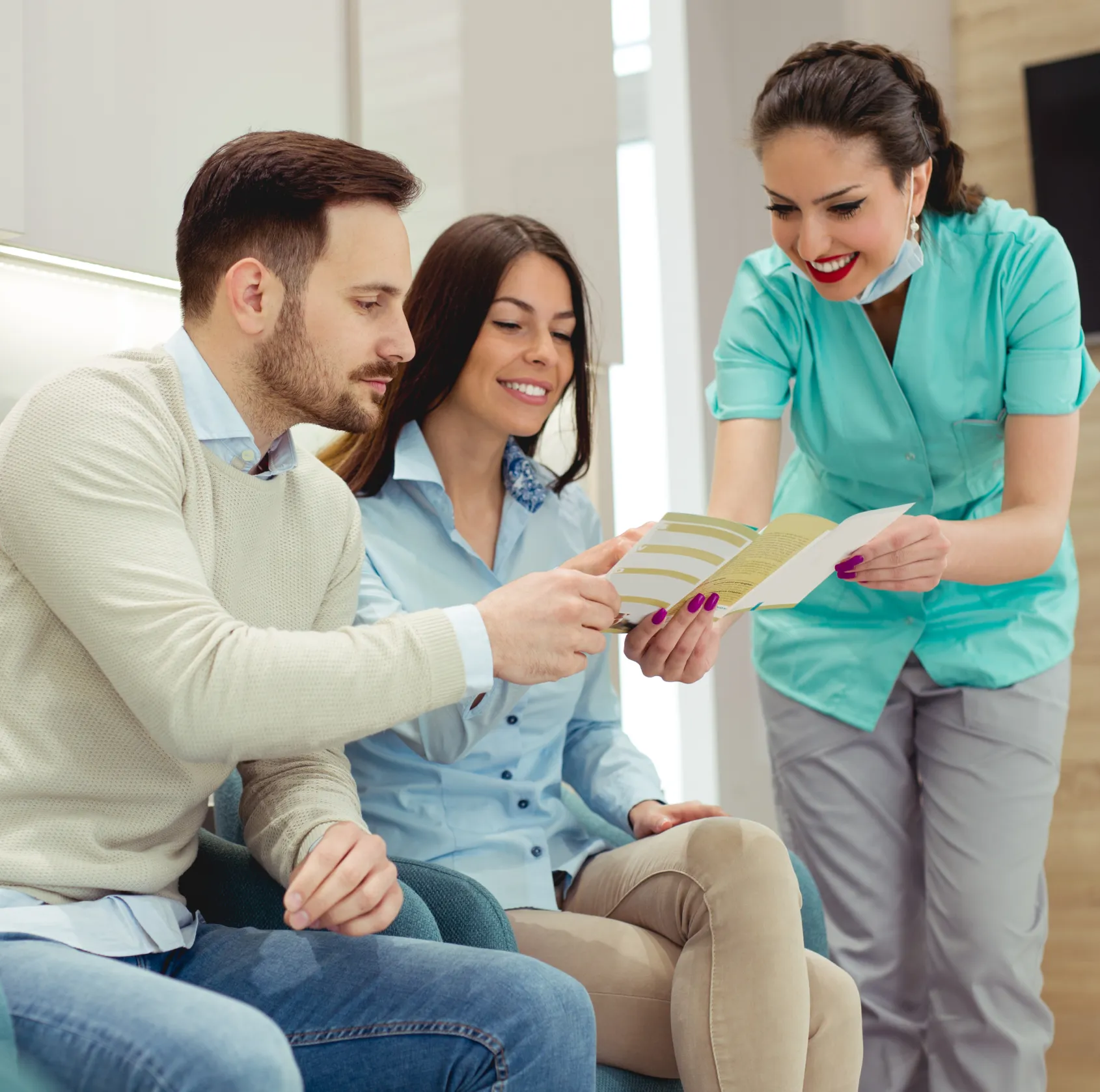 Smiling female healthcare worker in scrubs showing a brochure to a seated man and woman.