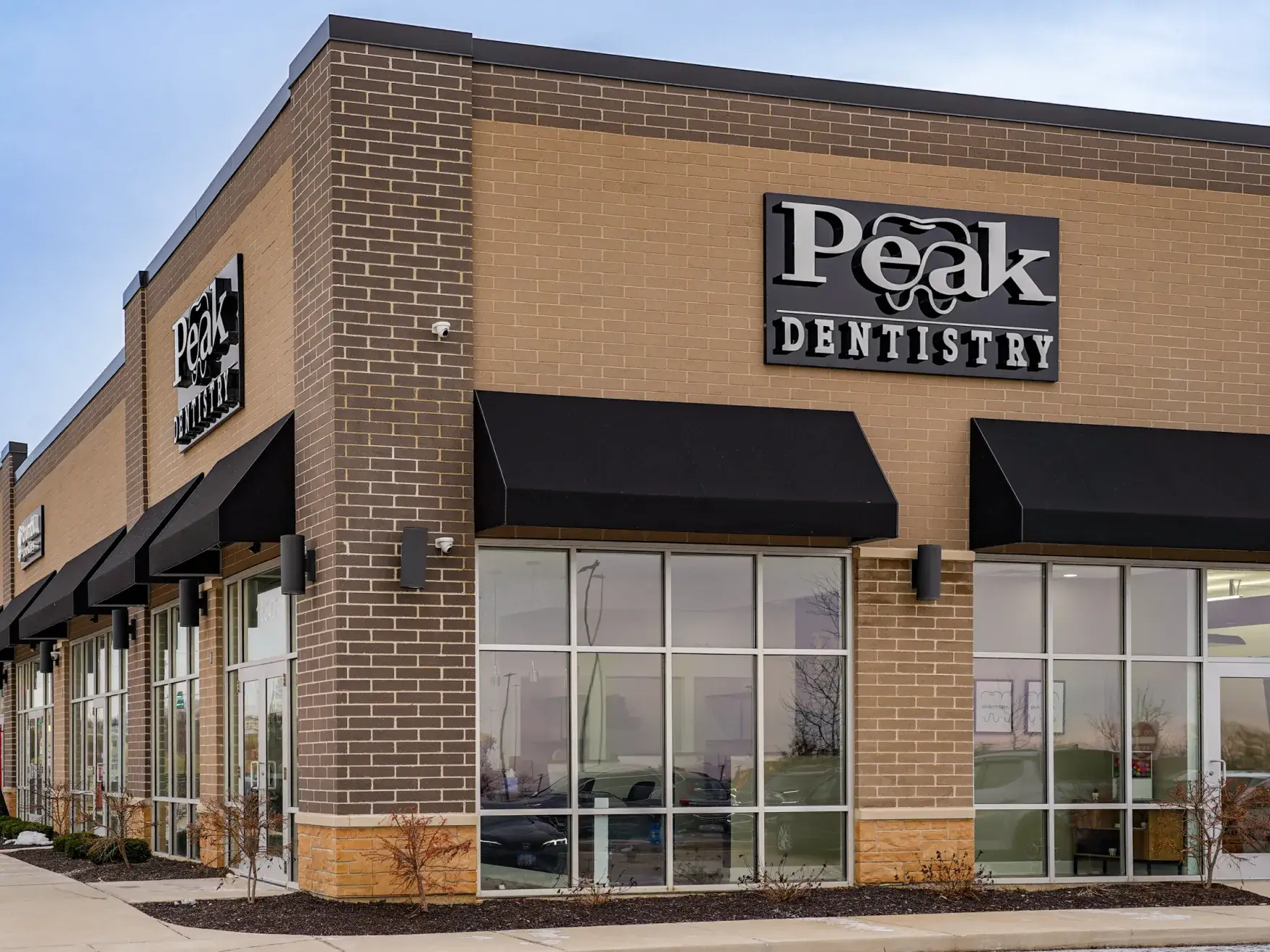 Exterior view of a brick building with large windows and black awnings, featuring a sign that reads 'Peak Dentistry'.