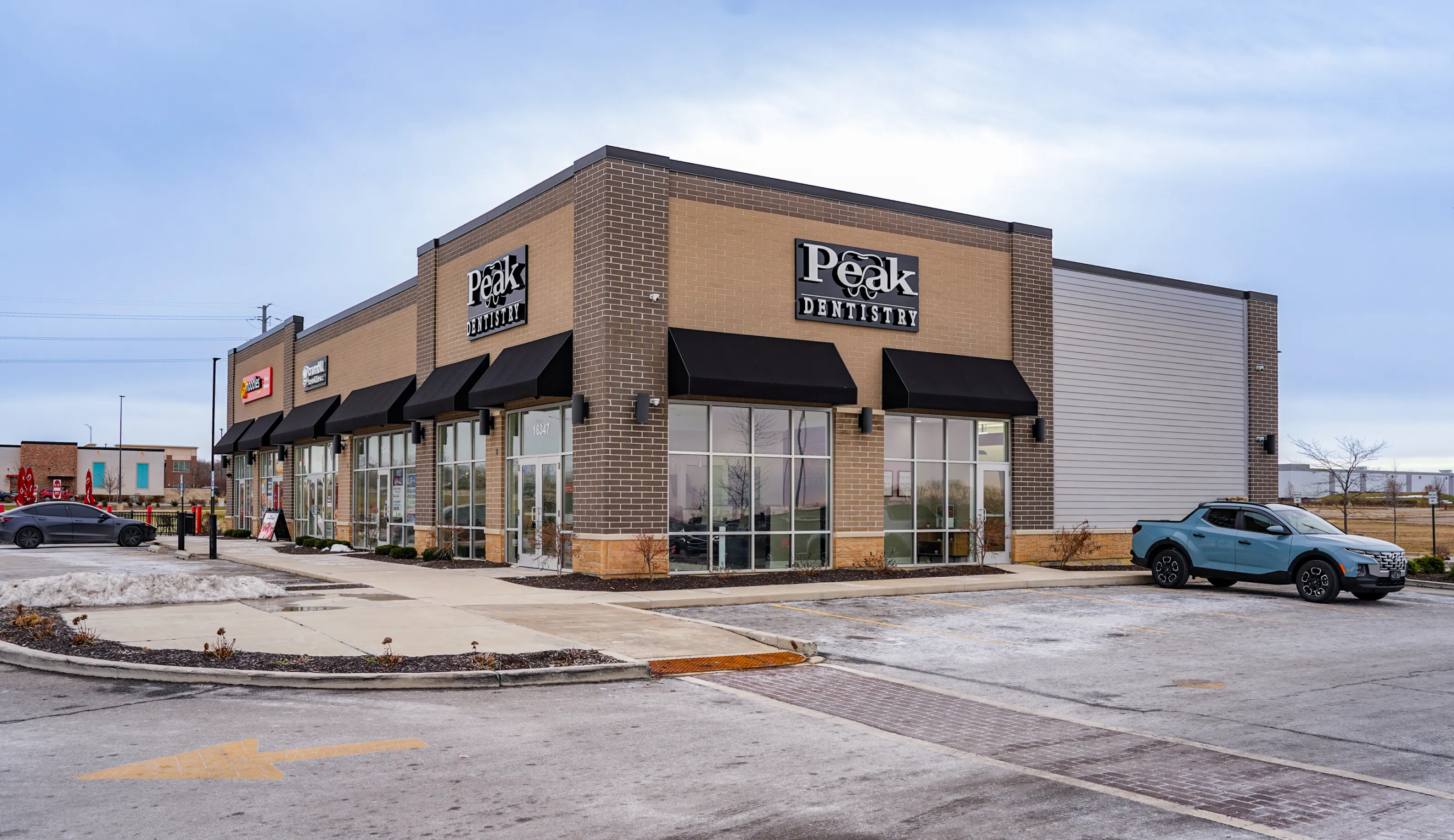 Corner view of a commercial building with a Peak Dentistry office and adjacent stores, with parked cars and a partly cloudy sky.