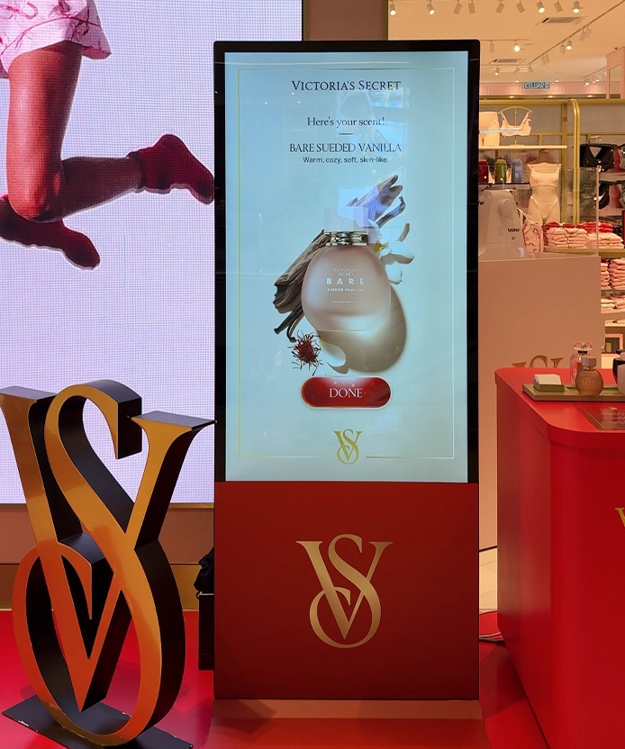 Victoria's Secret fragrance kiosk screen recommending Bare Sueded Vanilla beside a gold VS logo sculpture in-store