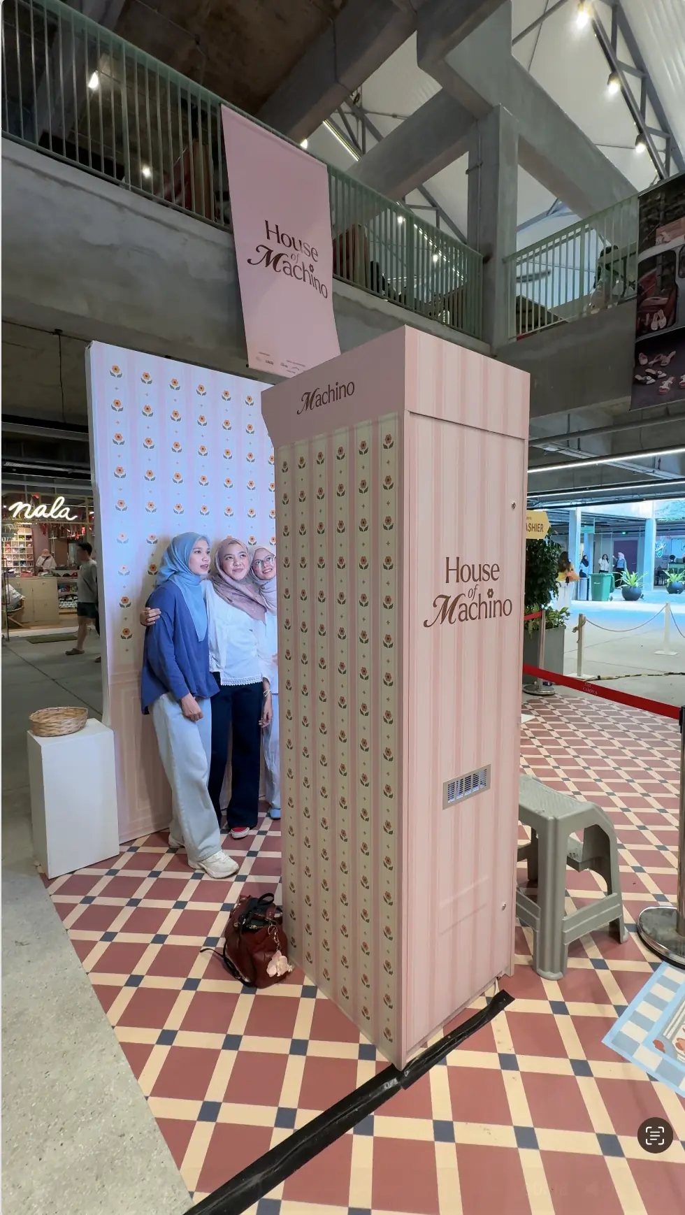 Three women posing together at the House of Machino floral backdrop next to the tall pink branded photo booth on checkered tile flooring