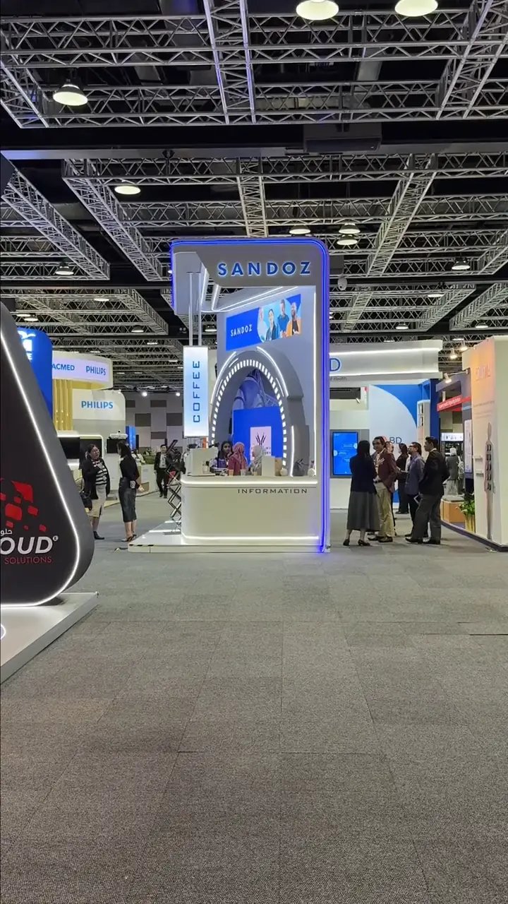 Sandoz exhibition stand with illuminated "Sandoz" signage, coffee and information counter, and attendees at the healthcare expo
