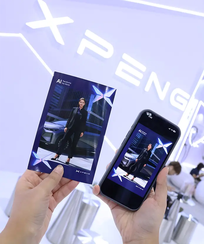 Printed XPENG AI photocard of a guest with an XPENG SUV held beside the digital copy on a phone