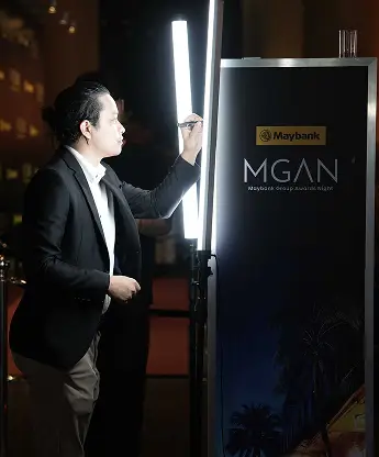 Guest in a suit signing the illuminated glass panel beside the Maybank MGAN branded standee