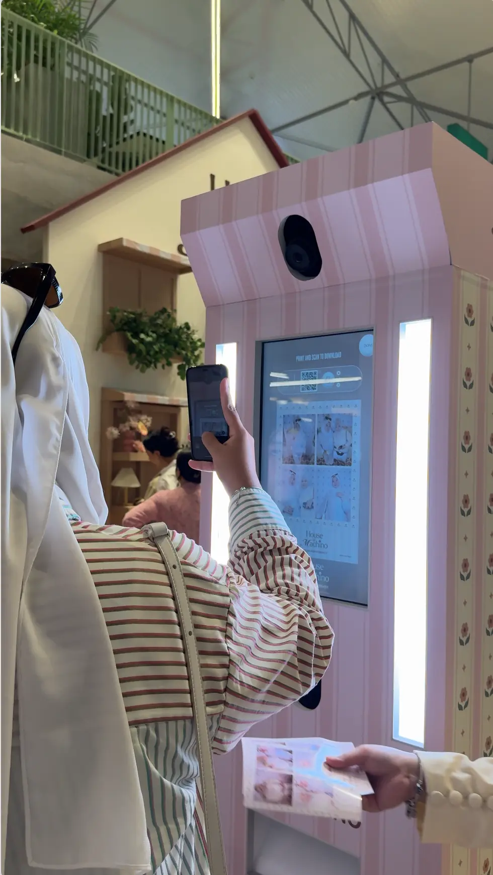 Shopper scanning the House of Machino photo booth's QR code with her phone to download photos, holding a printed strip in the other hand