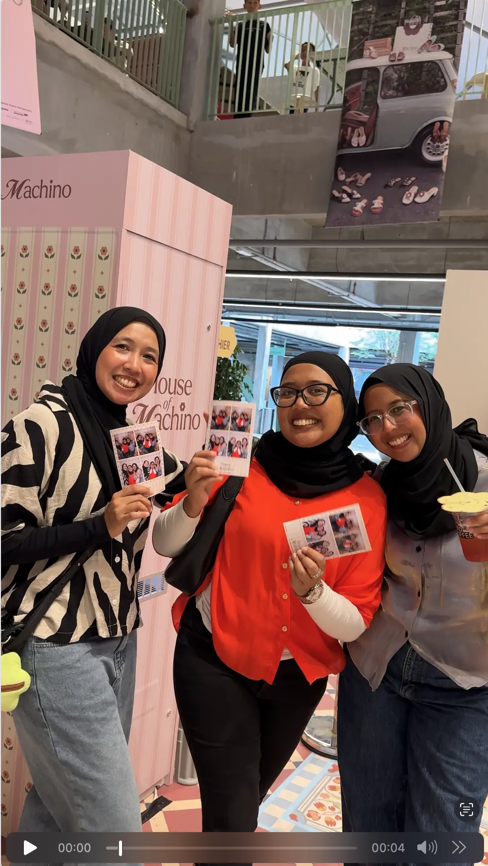 Three friends in hijabs smiling and holding their printed House of Machino photo strips beside the pink booth at the pop-up event