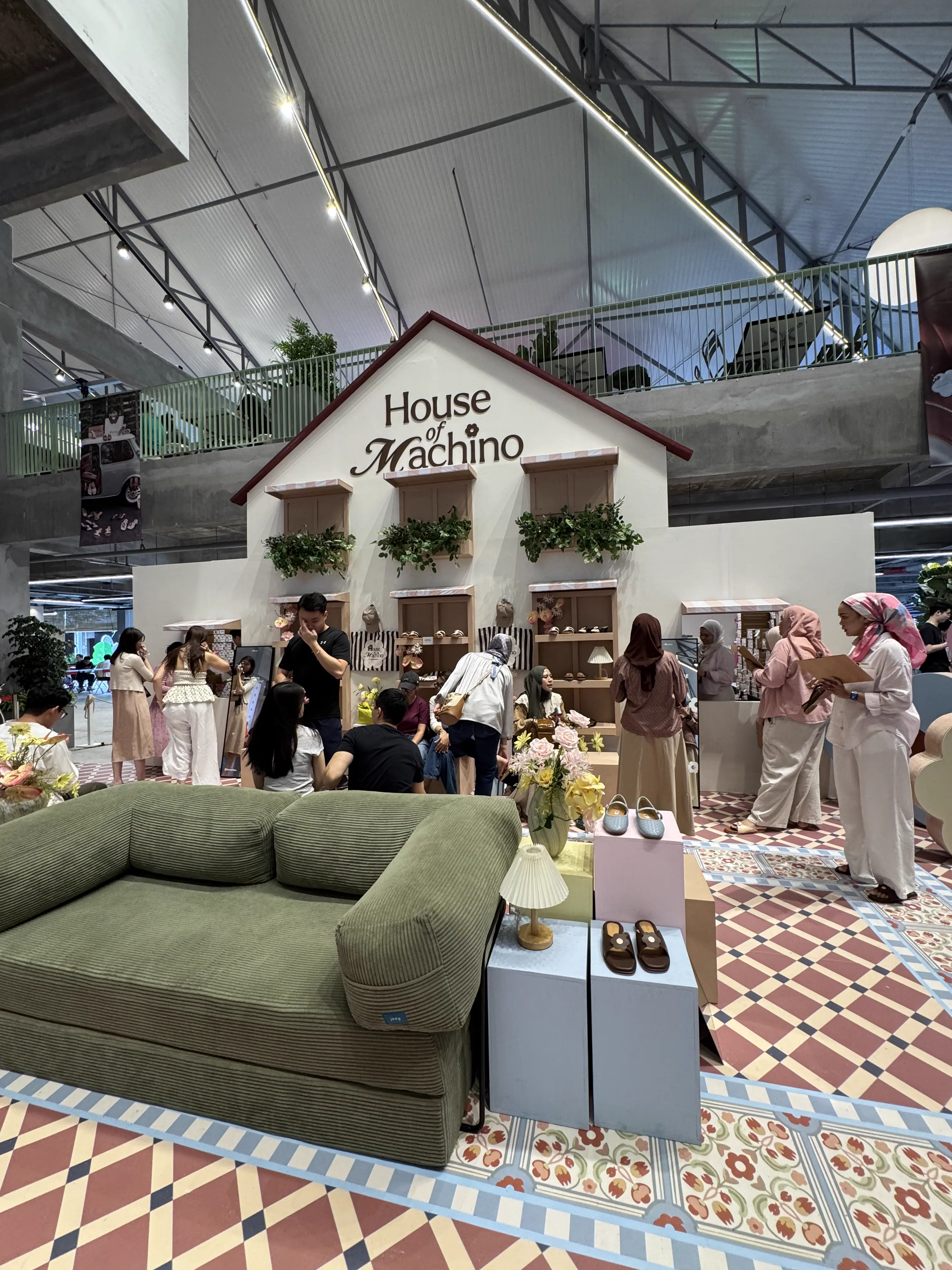 House of Machino pop-up storefront with a pitched-roof facade, footwear displays, a green corduroy sofa and shoppers browsing on decorative tiled flooring