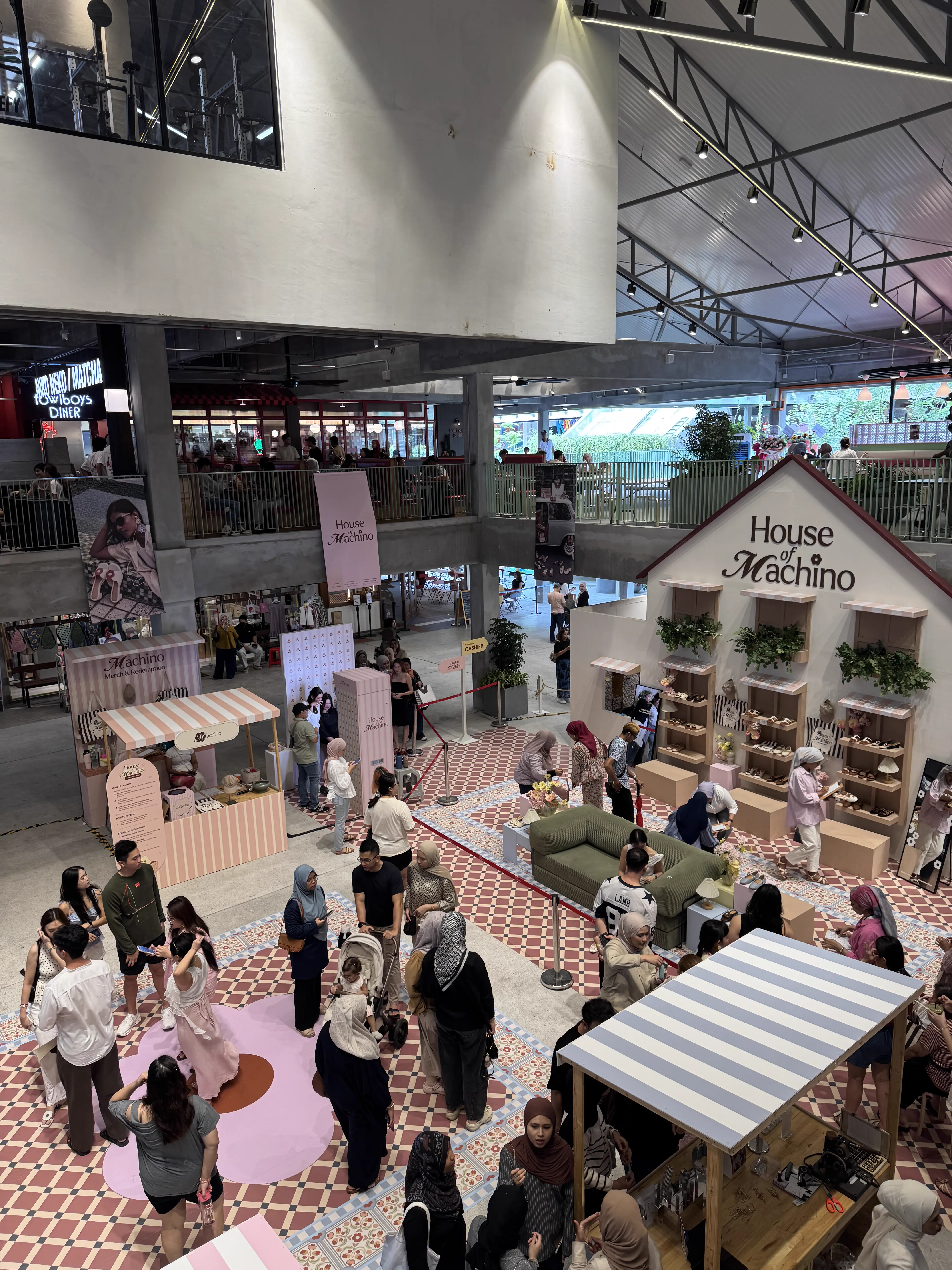 Aerial view of the House of Machino pop-up event in a mall atrium, with crowds browsing the house-shaped booth, striped kiosks and patterned tile flooring