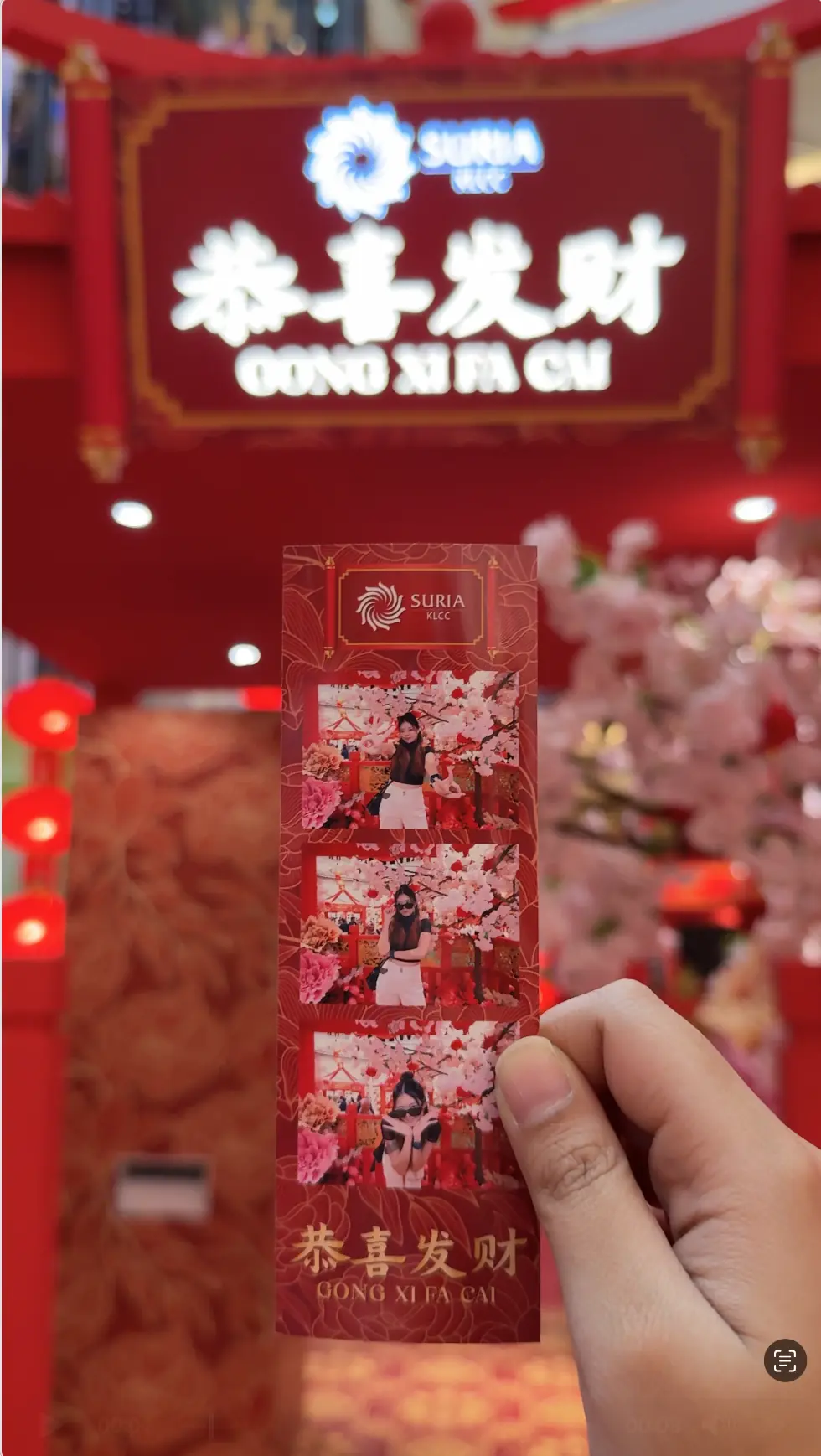 Hand holding a printed Suria KLCC Gong Xi Fa Cai photo strip in Kuala Lumpur, beside cherry blossom decorations