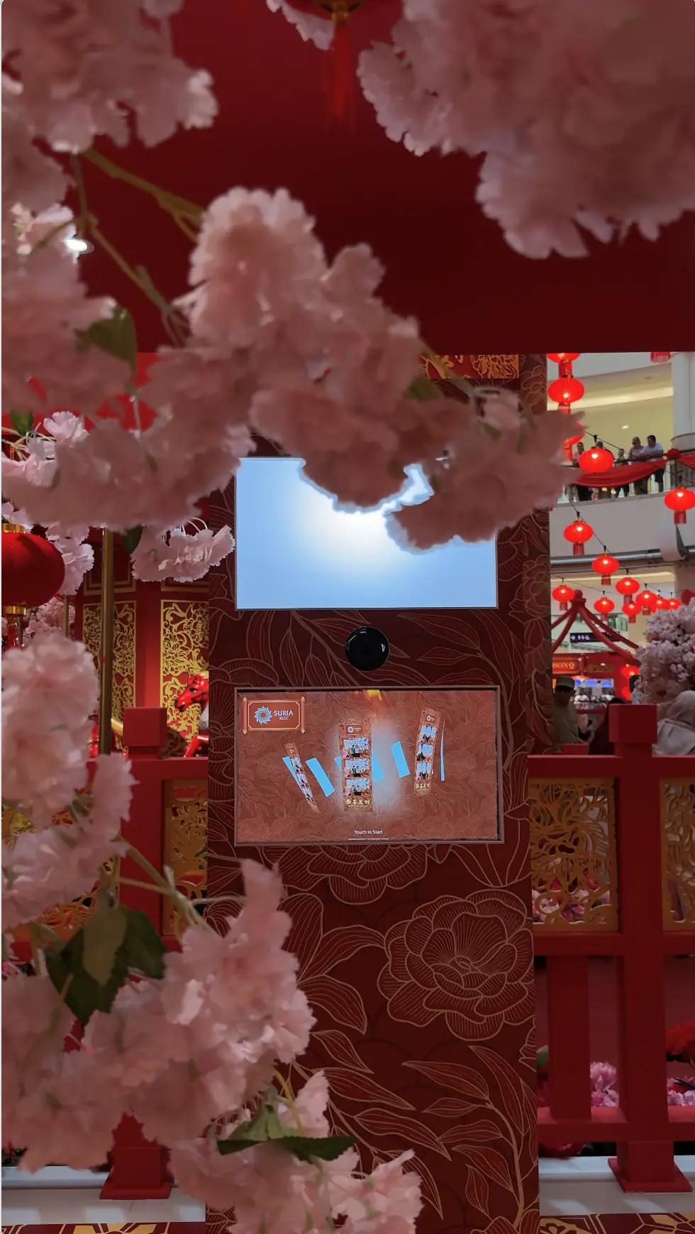Suria KLCC Chinese New Year photo booth framed by pink cherry blossoms, red lanterns and a touchscreen displaying the "Touch to Start" welcome screen