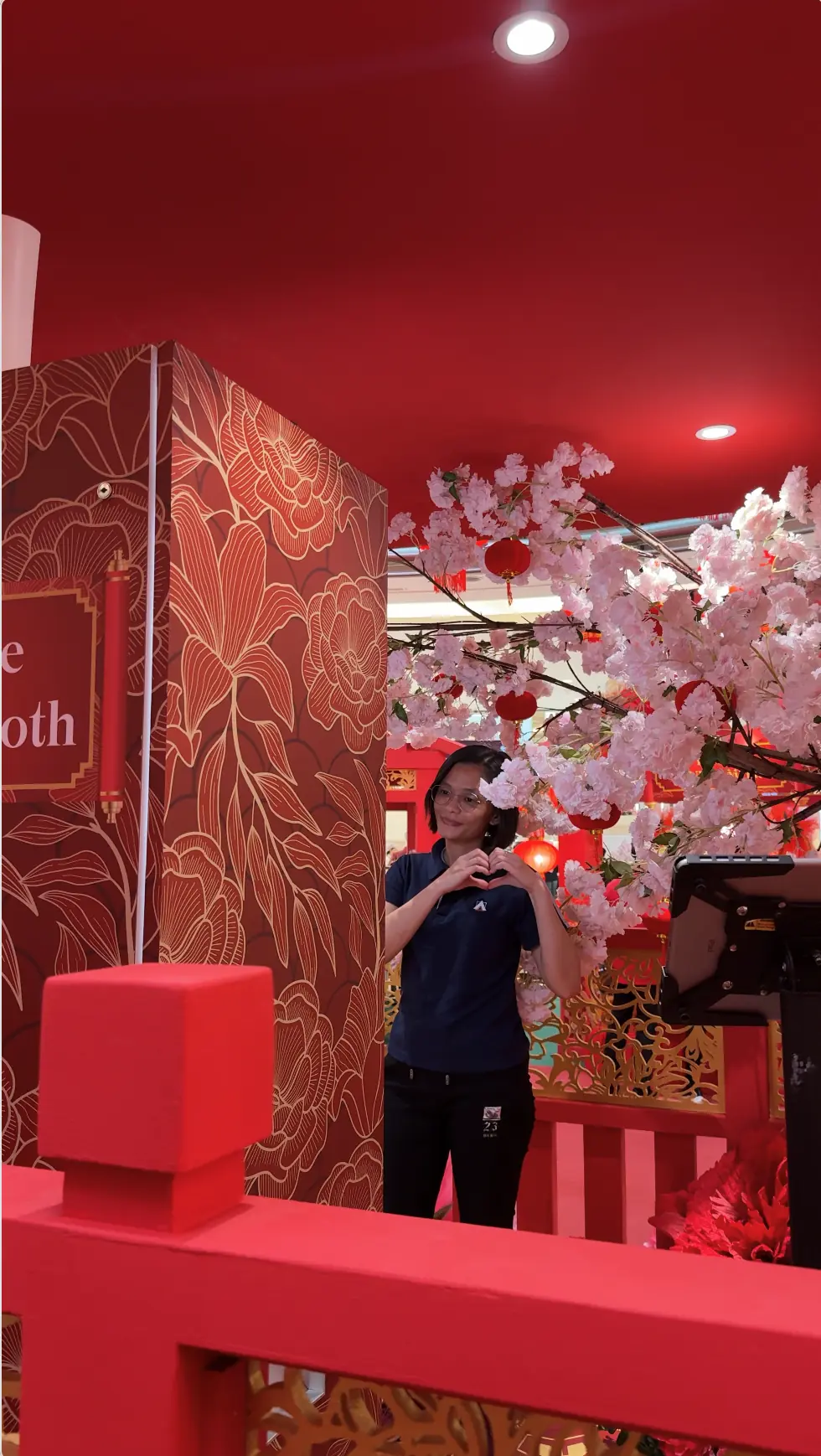 Suria KLCC staff member making a heart gesture beside the red floral photo booth and cherry blossom tree at the Chinese New Year setup