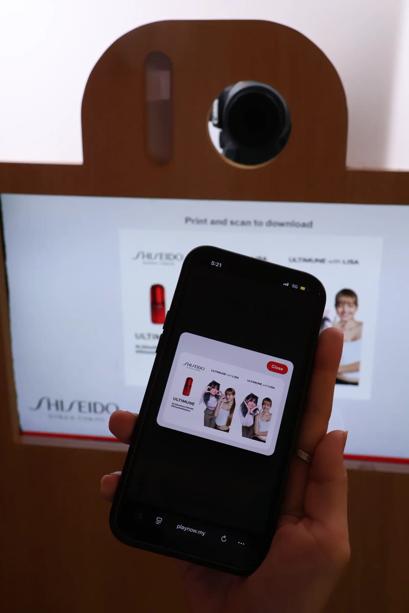 Phone displaying a downloaded ULTIMUNE x LISA photo strip in front of the booth's print-and-scan screen at the Shiseido pop-up