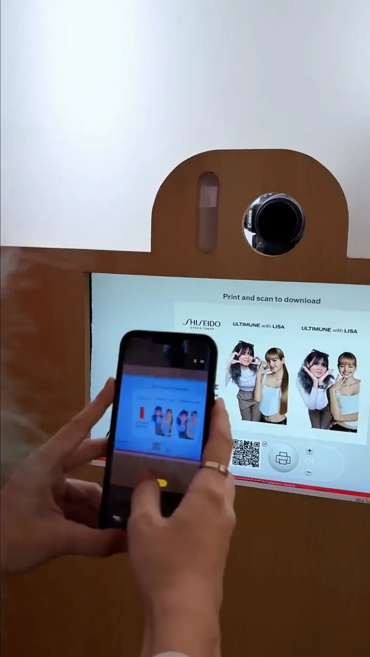 Visitor photographing the ULTIMUNE x LISA photo strips on the booth's print-and-scan screen with a phone