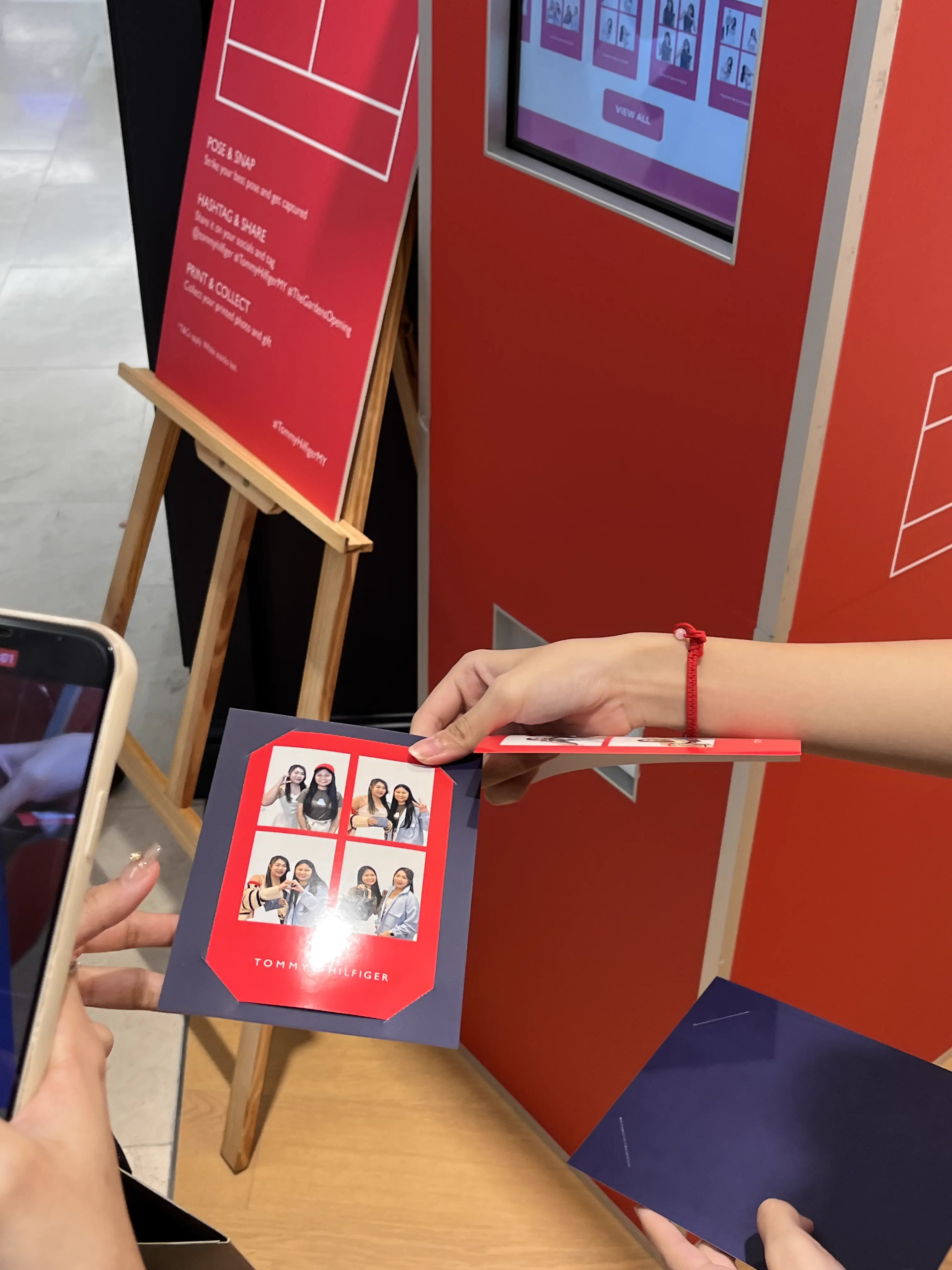 Hand holding a Tommy Hilfiger branded grid photo strip beside the booth's instructions easel and kiosk screen