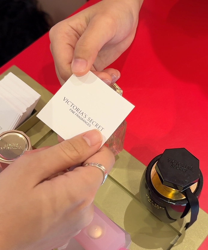 Hands holding a Victoria's Secret Fine Fragrances scent card over gift boxes and a perfume bottle at the counter