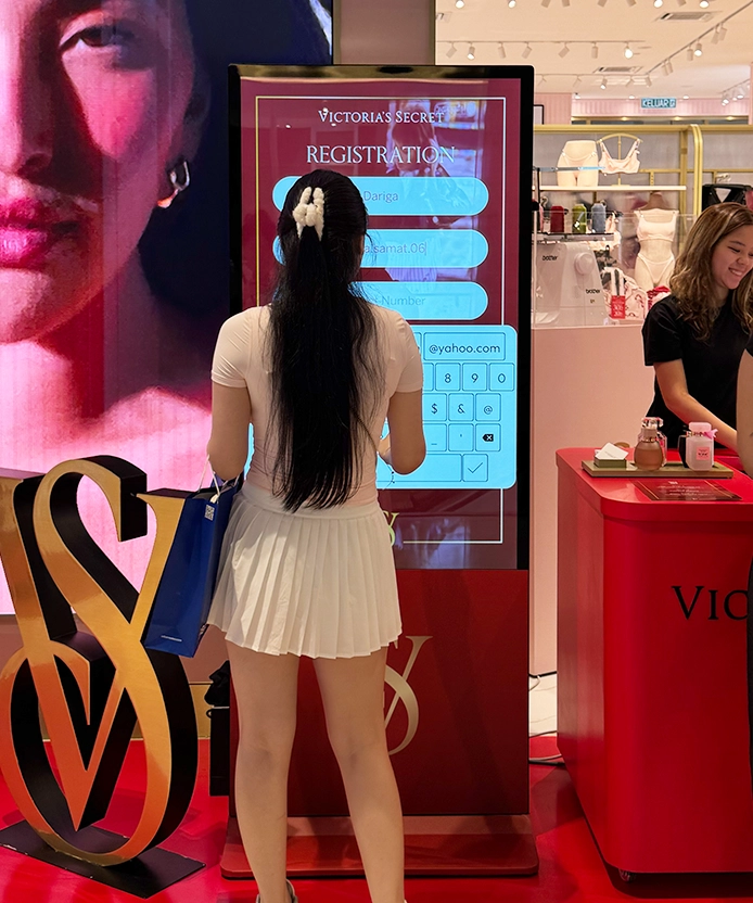 Customer entering her details on the Victoria's Secret registration touch screen beside a gold VS logo display