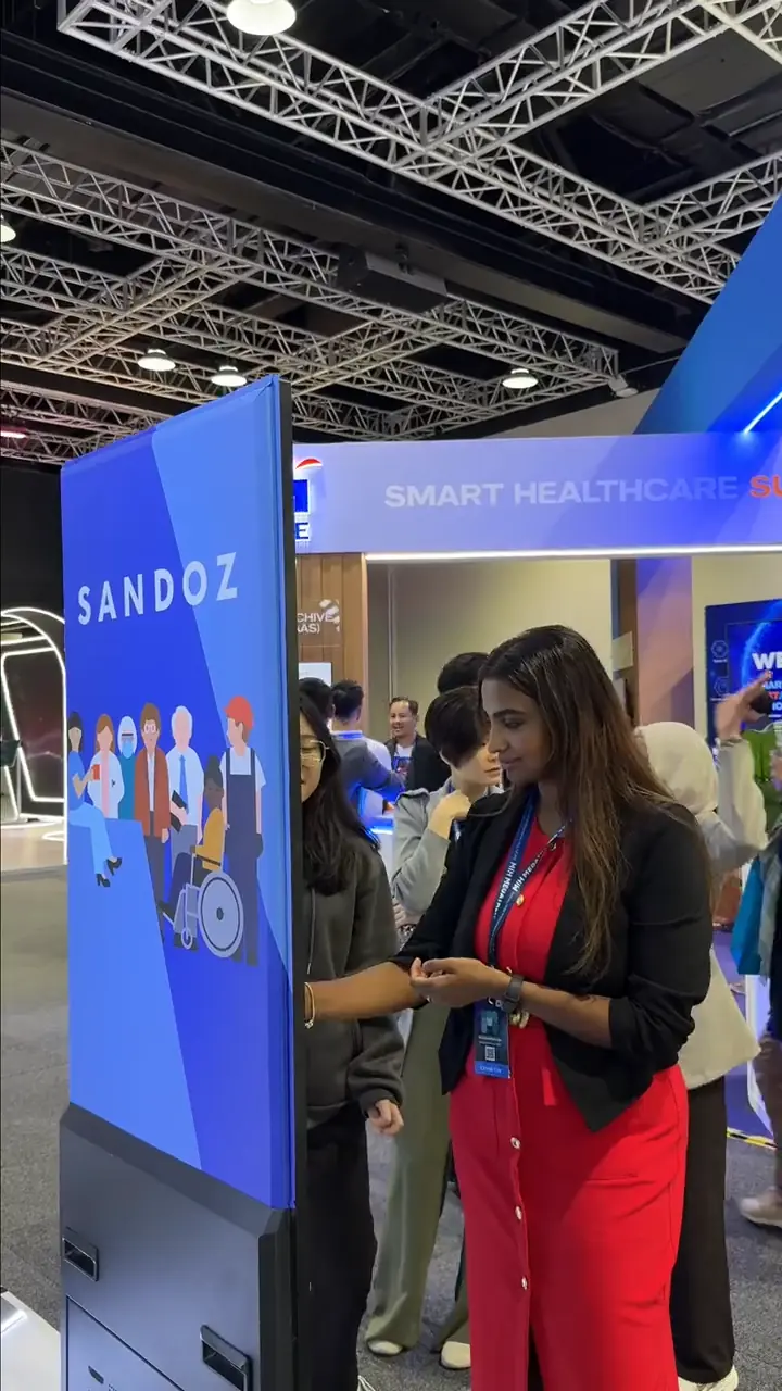 Sandoz brand ambassador in a red outfit standing beside the booth's tall display screen at the Smart Healthcare expo